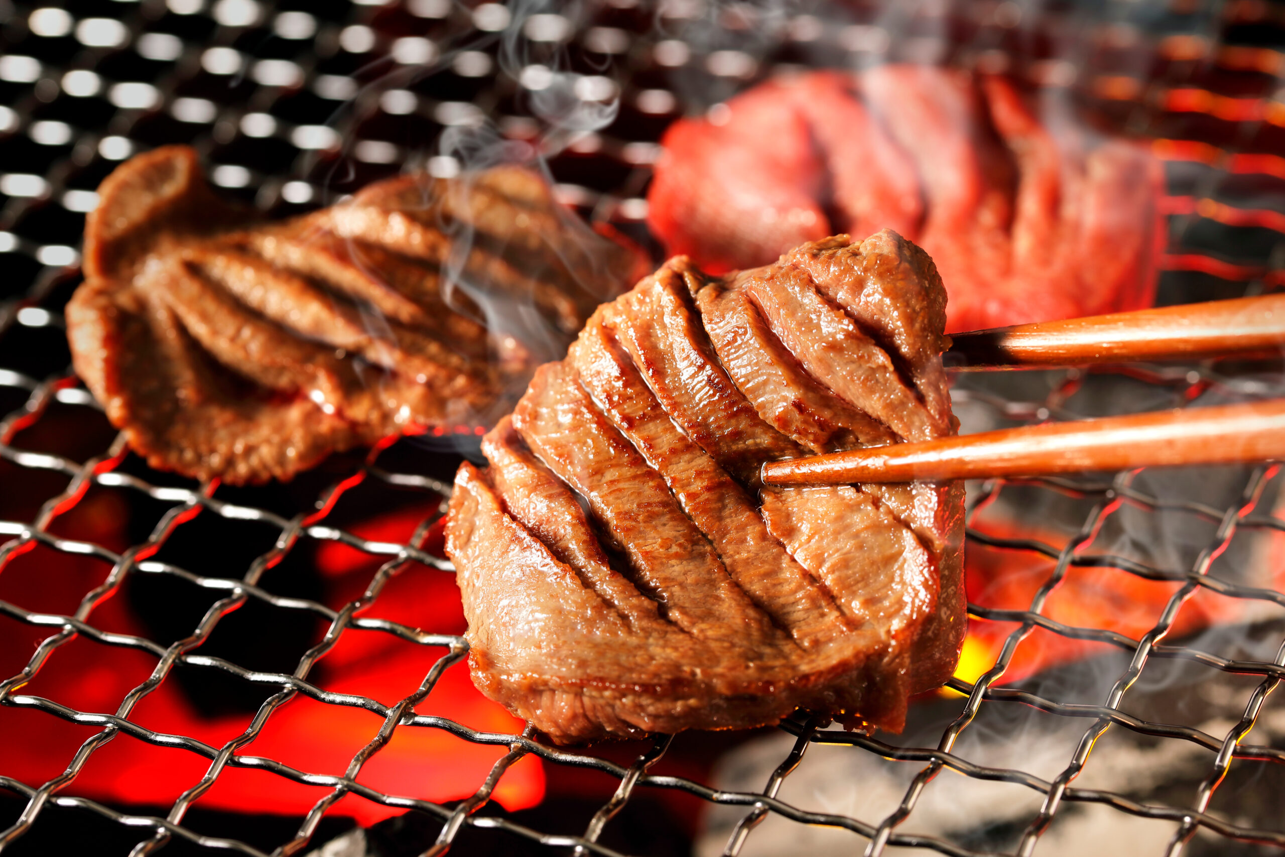 Beef steak pieces grilling on a wire grate over hot coals with wispy smoke.