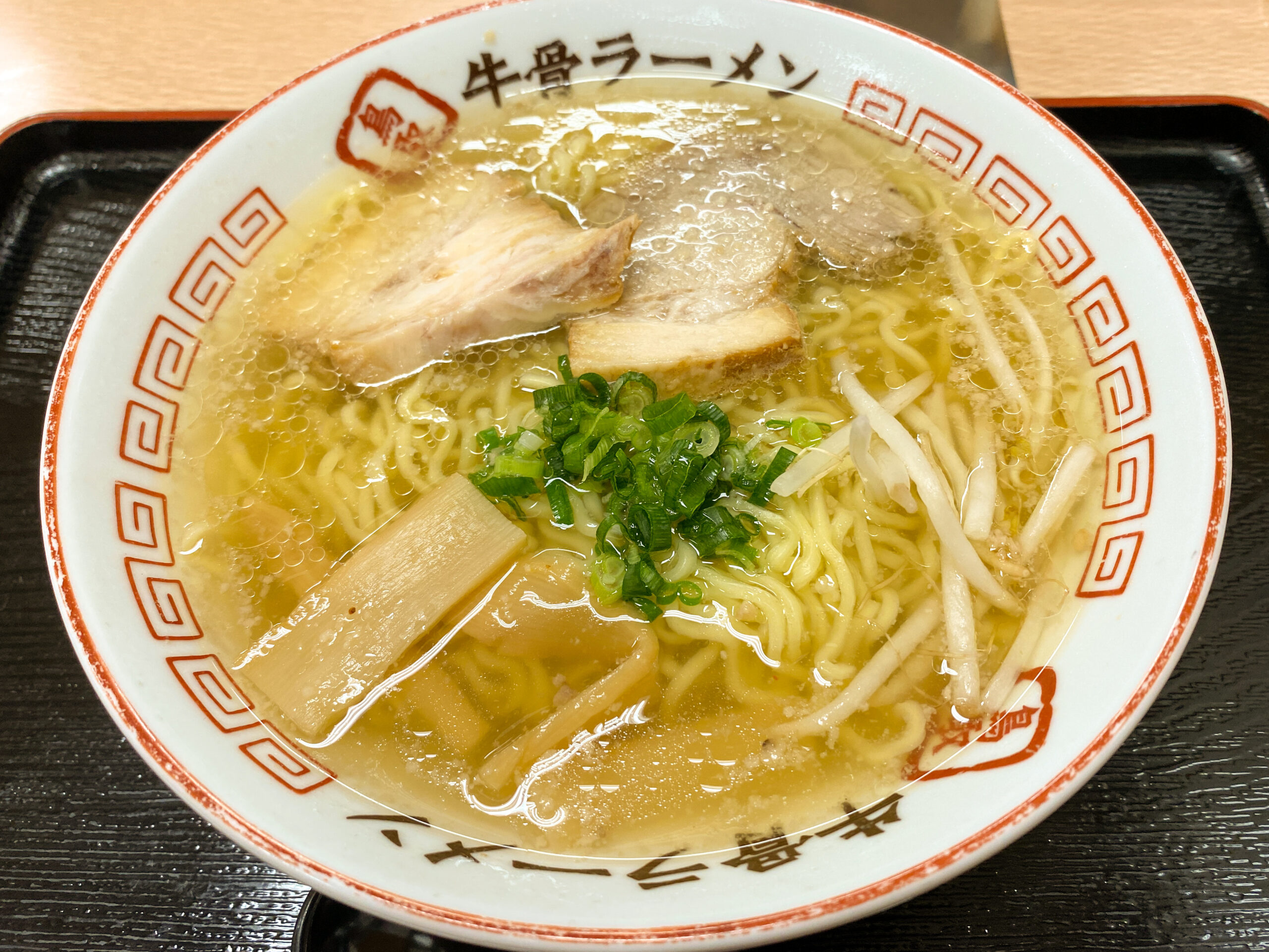Ramen bowl with sliced pork, green onions, and bamboo shoots in a clear broth on a dark table.