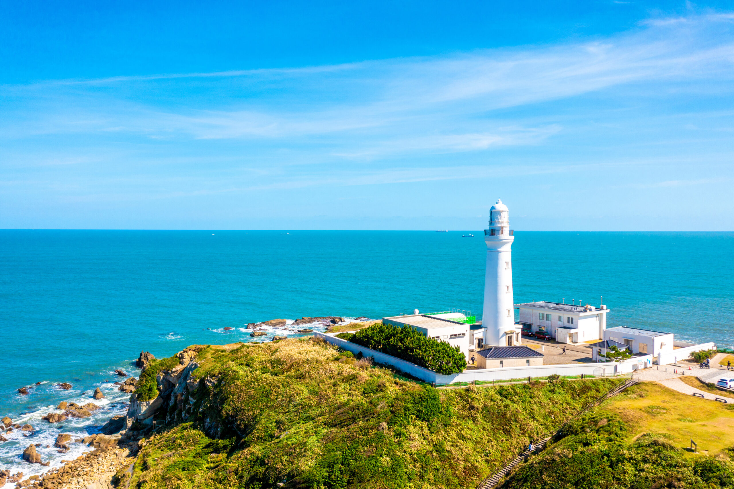 White lighthouse on a rocky coastline with blue ocean and clear sky in the background.