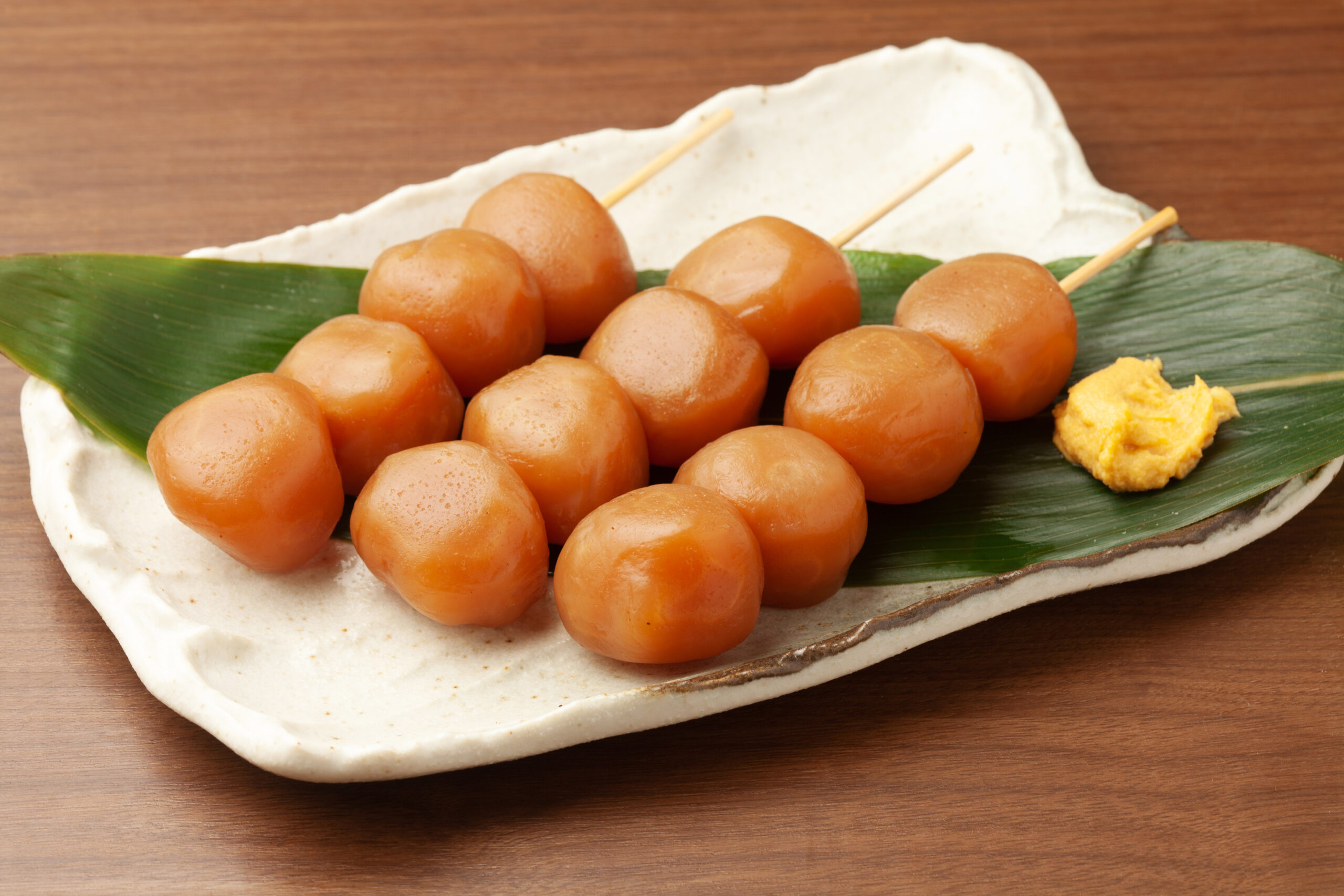 Skewered dango dumplings on a leaf‑lined plate with a dab of yellow paste beside them, close view.