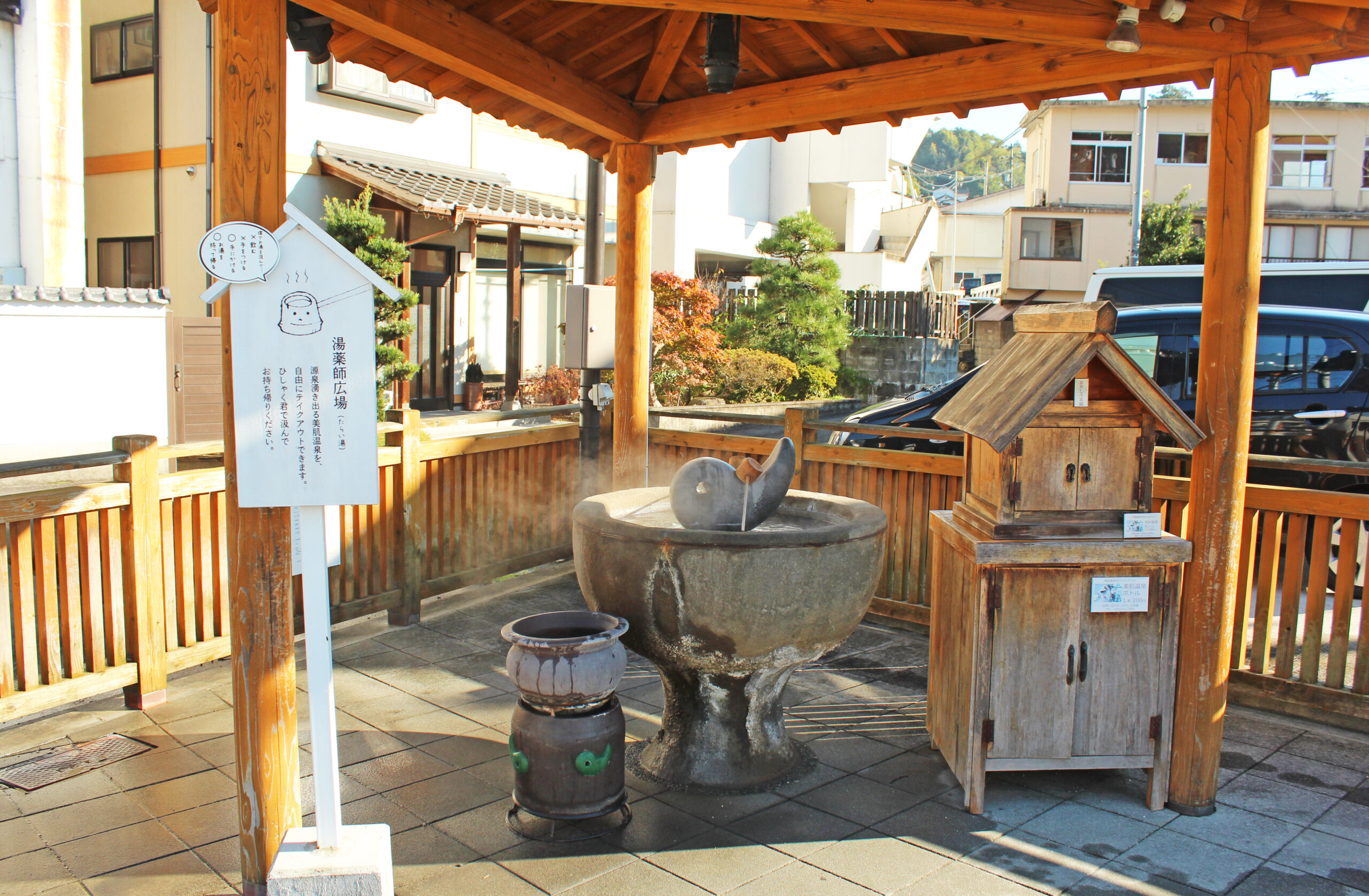 Wooden pavilion over a stone courtyard with a large round water basin, decorative spout sculpture, and a carved wooden well, set against modern buildings in the background.