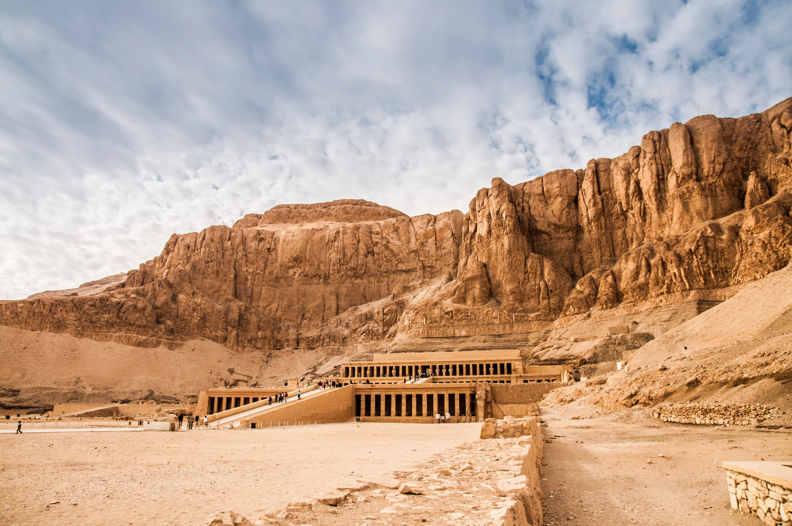 Desert scene with a monumental temple built into red cliffs and visitors walking up a ramp toward the entrance.