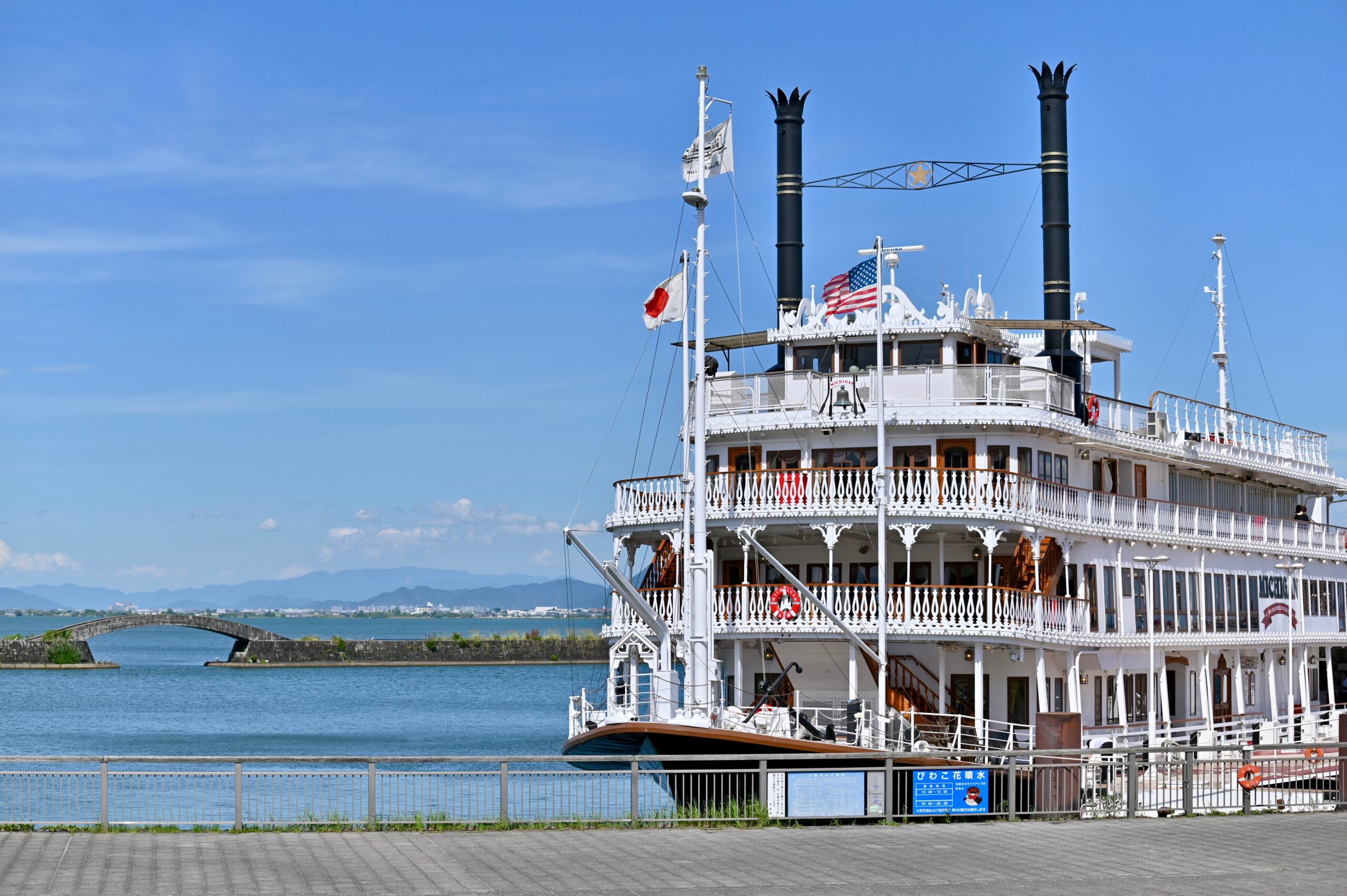 Ornate white riverboat with two black smokestacks docked by the water under a clear blue sky.