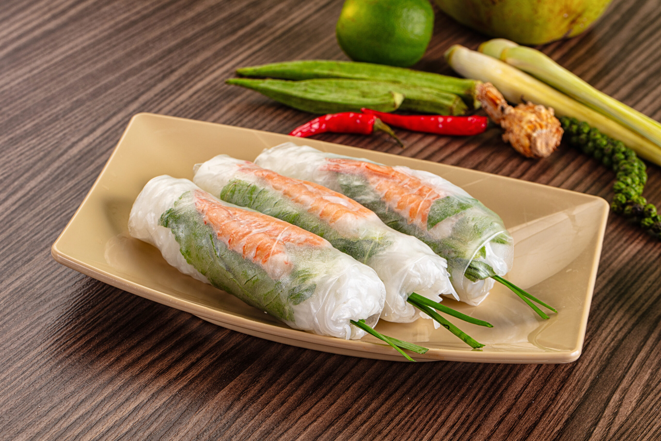 Fresh rice-paper spring rolls filled with vegetables on a beige plate, with green herbs and veggies in the background on a wooden table.