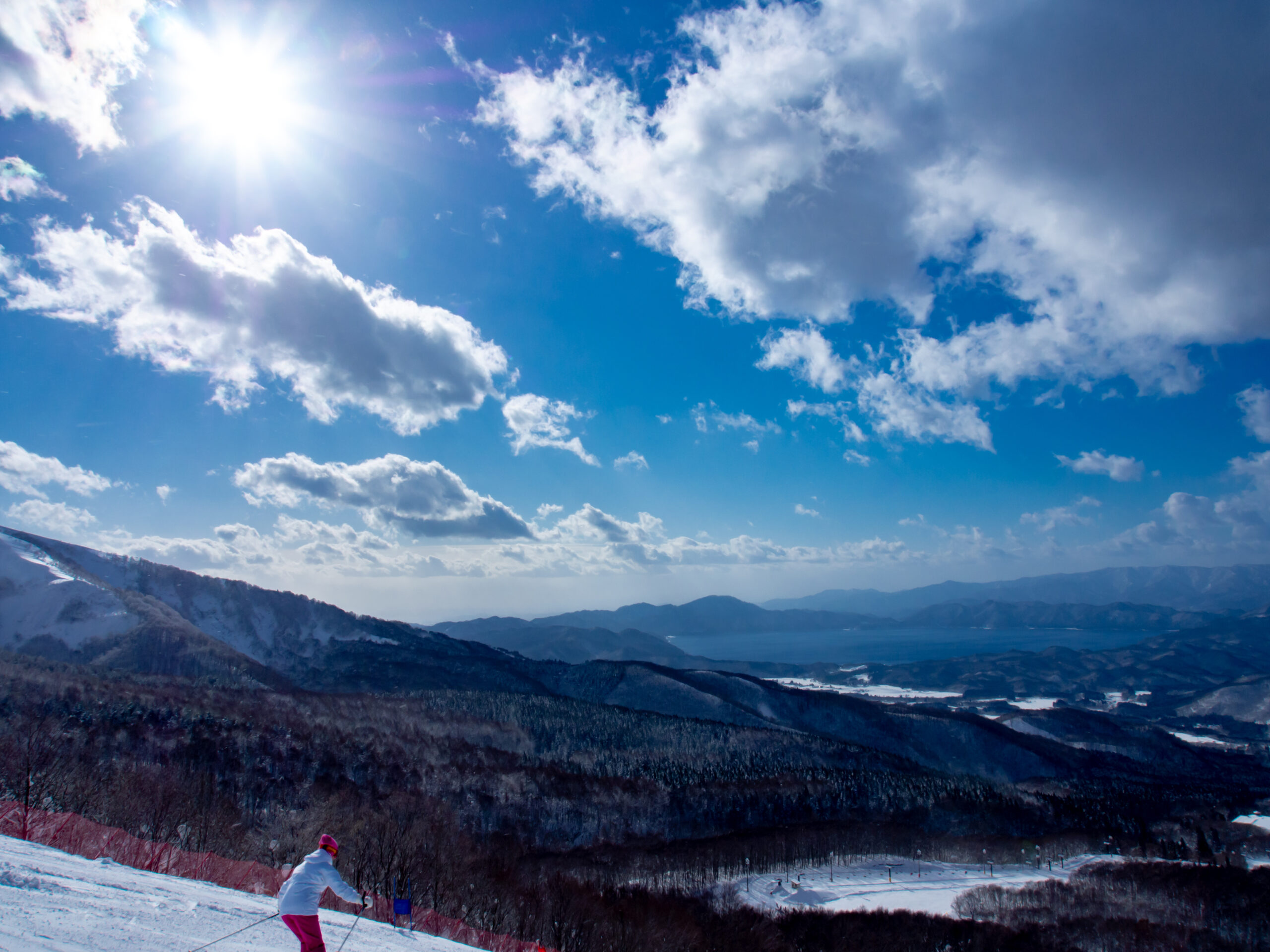 Snowy mountain slope under a bright sun with blue sky and clouds; skier in white jacket and pink pants near the bottom left.