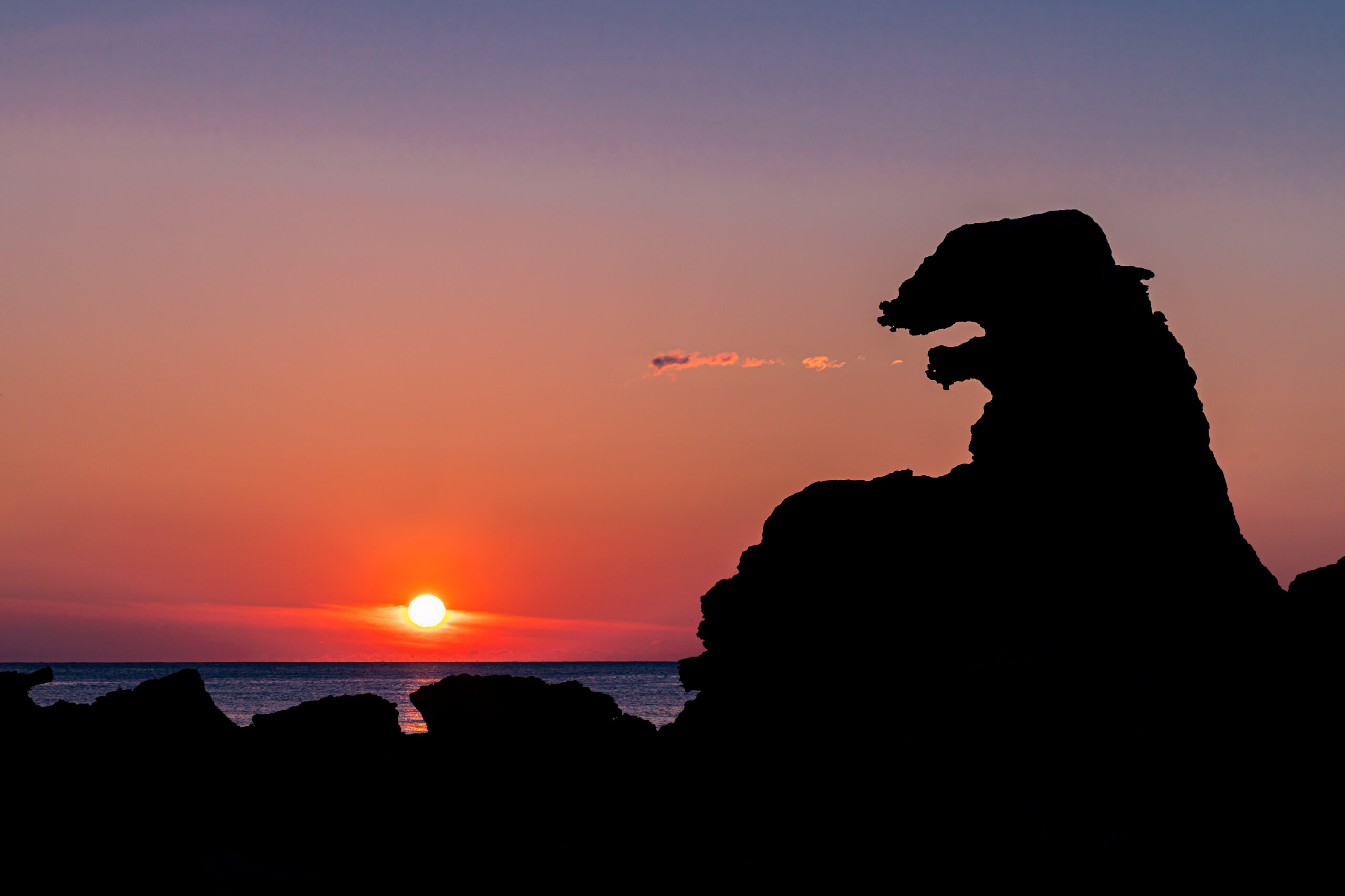 Silhouetted jagged rock formation against a sunset sky with orange-pink hues over the sea.