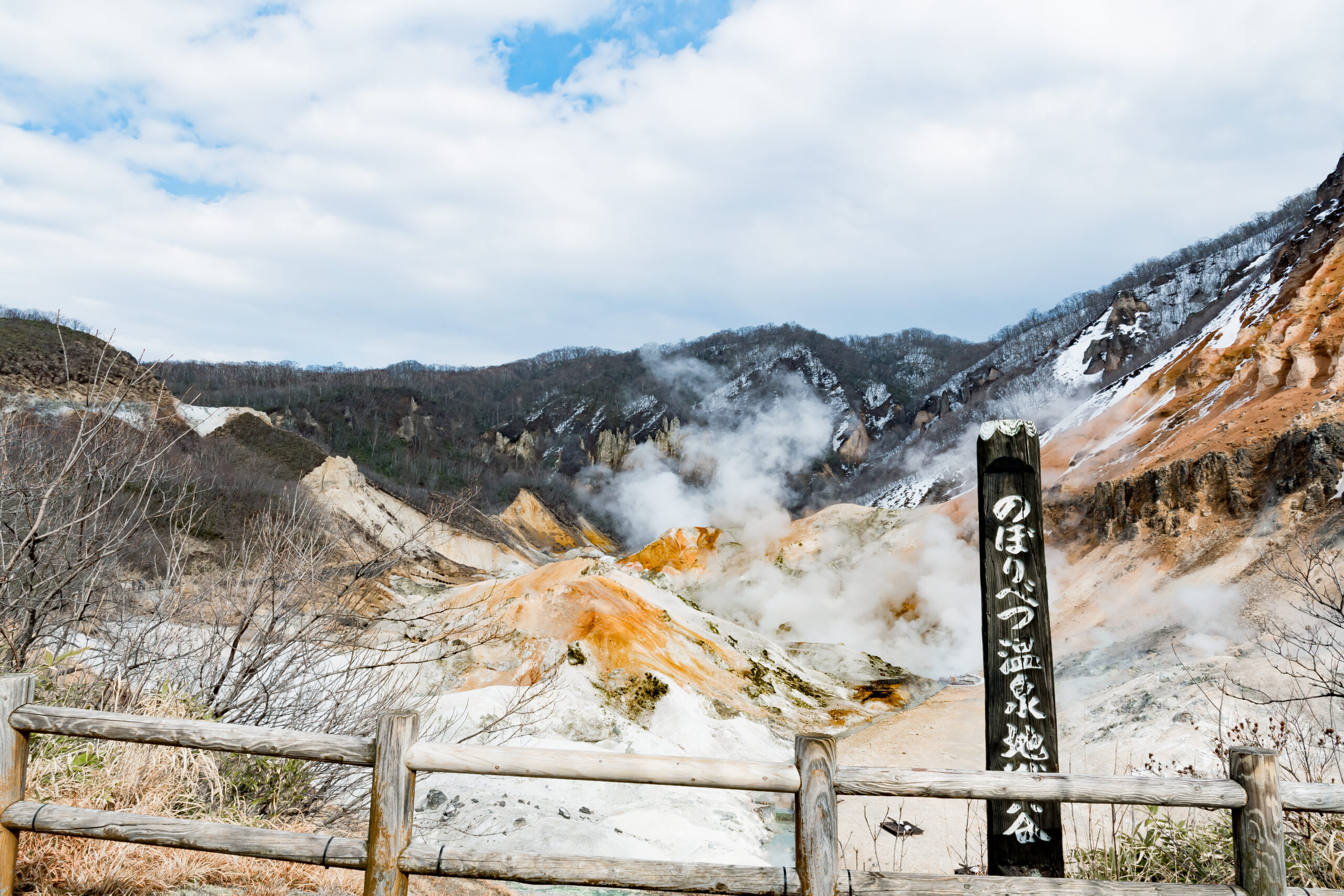 Outdoor geothermal area with steam rising from orange and yellow rocks, a wooden railing, and a tall sign with Japanese writing.