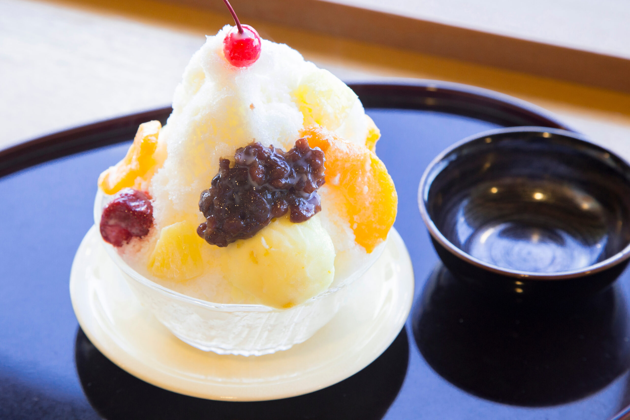 Shaved ice dessert topped with a red cherry and yellow fruit pieces in a glass, on a dark tray and plate.
