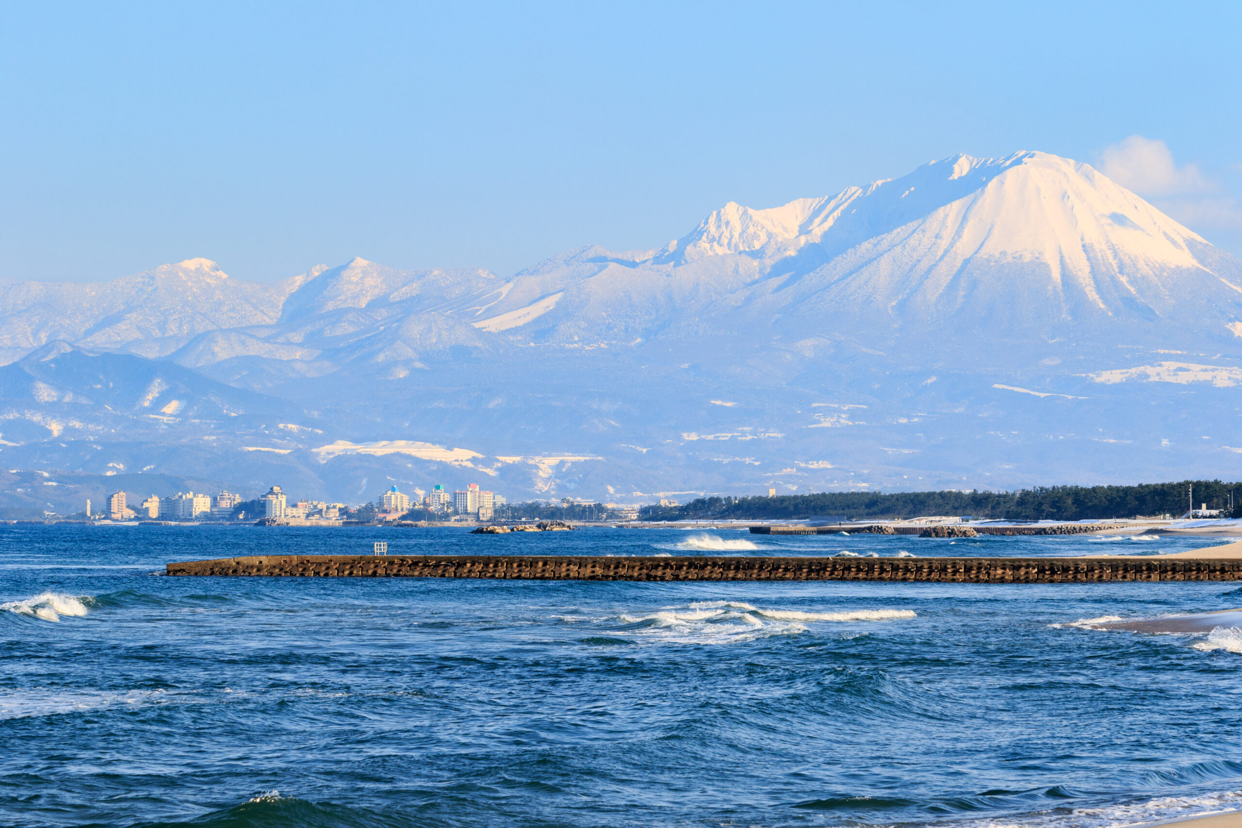 Coastal town at the shore with a snow‑capped mountain range rising behind and a clear blue sky above.