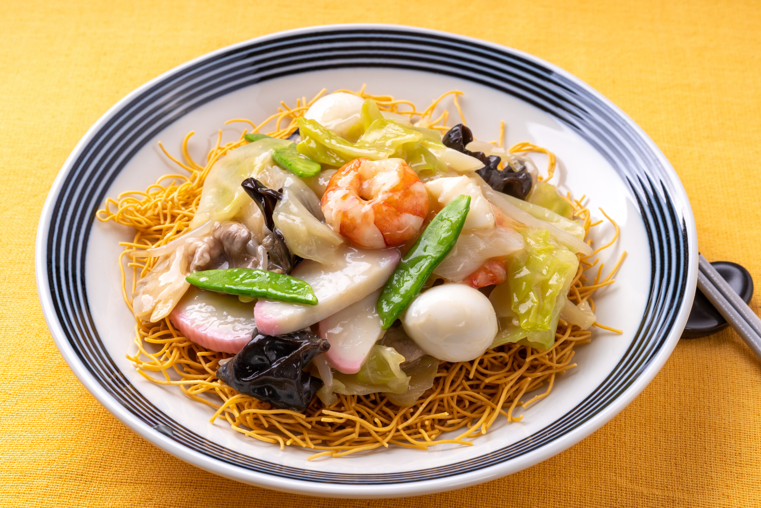Plate of crispy fried noodles topped with shrimp and assorted vegetables in a glossy sauce on a white plate with blue rim.