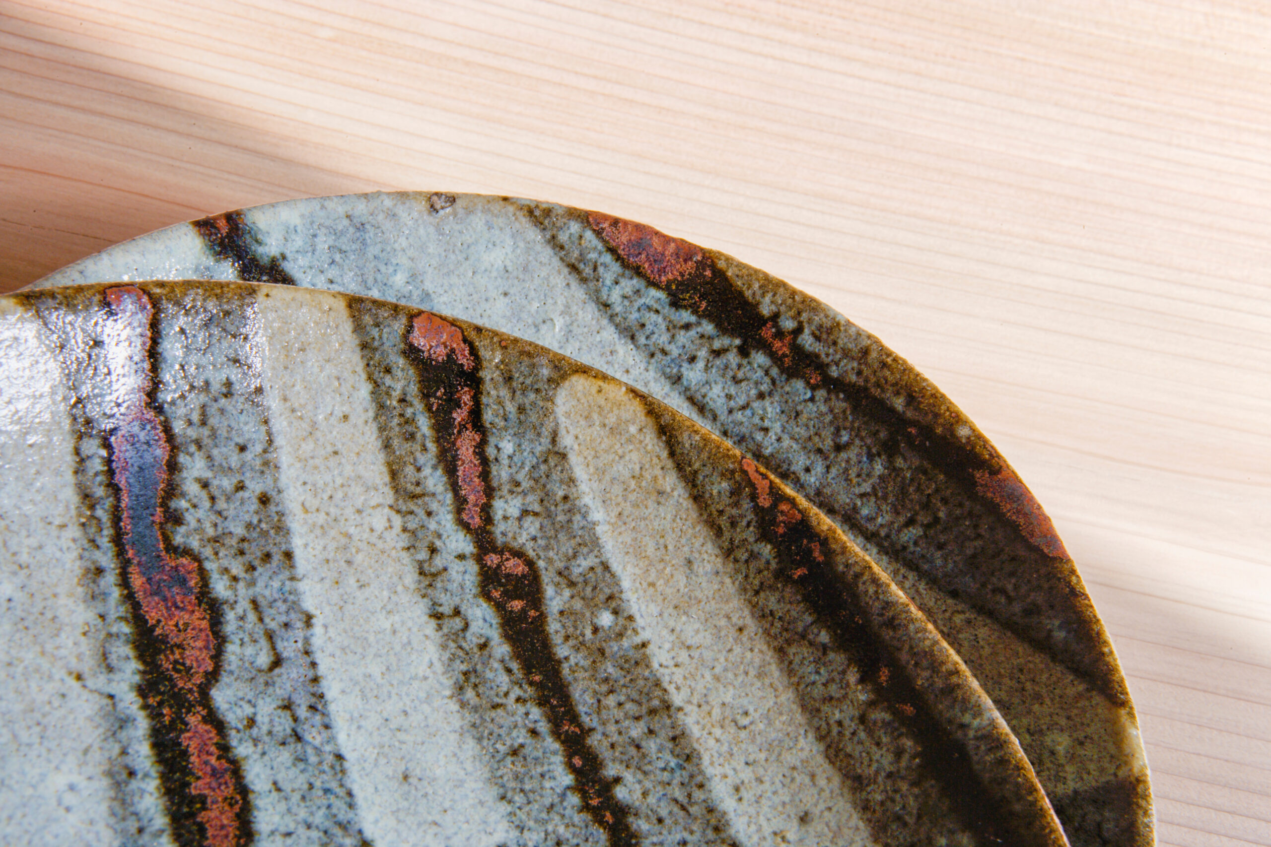 Close-up of a ceramic plate with blue glaze and rust-colored streaks on a pale wooden surface.