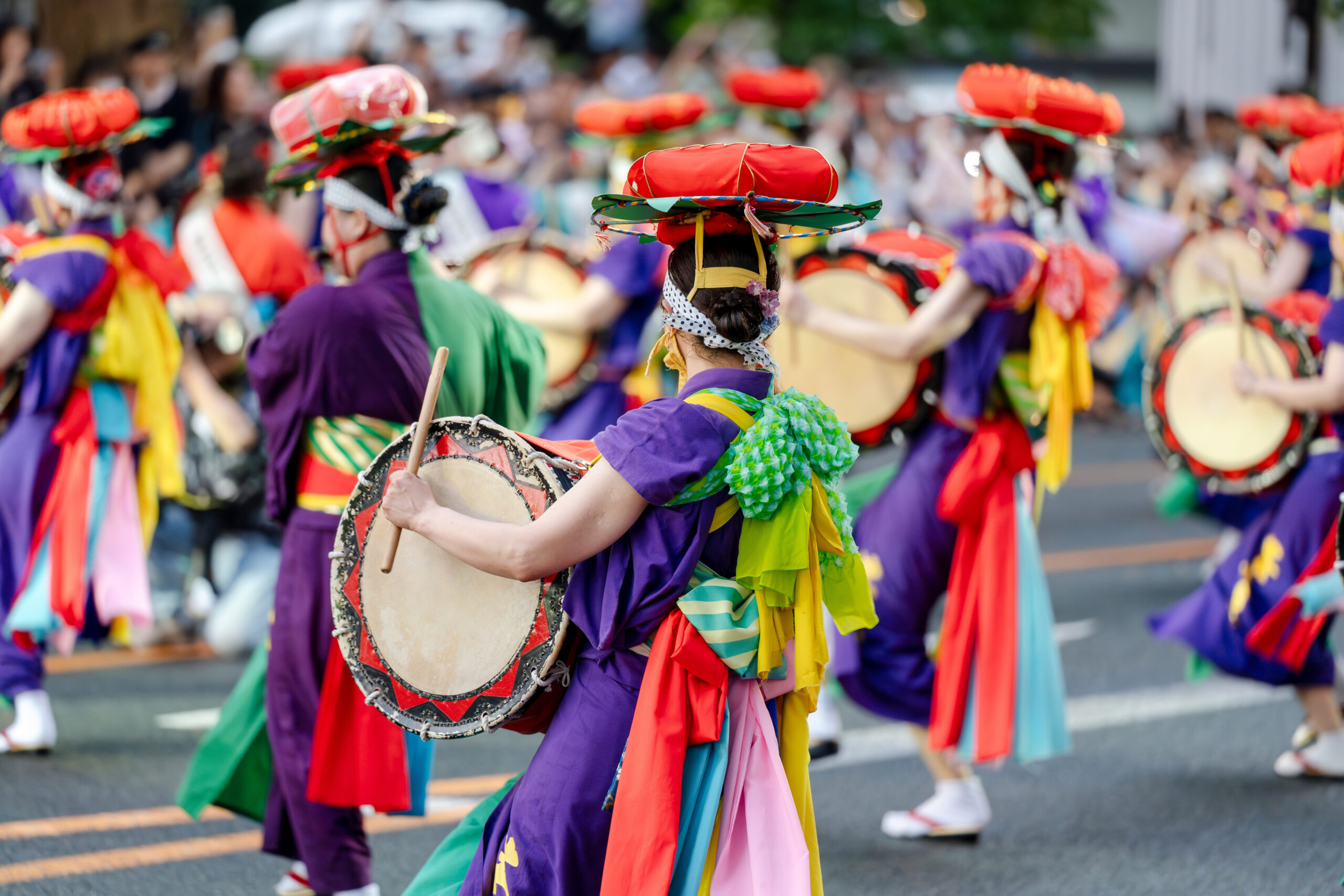 Colorful parade dancers in bright costumes playing drums on a city street, center performer with red headpiece.