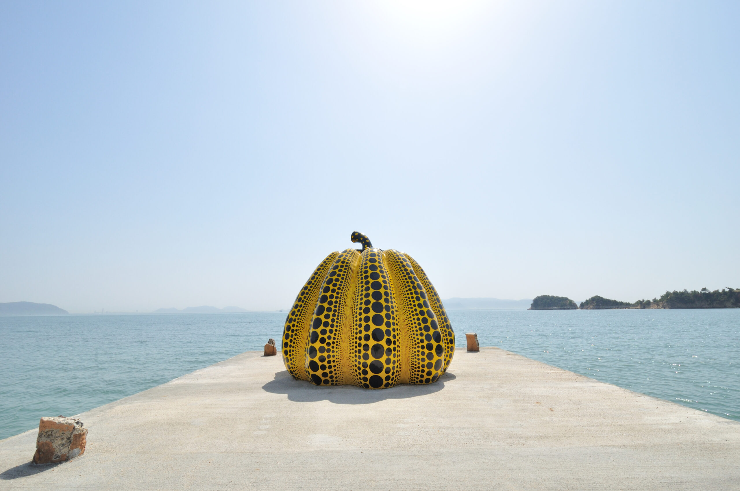 Large yellow sculpture with black polka dots sits on a concrete pier by a calm blue sea under a clear sky, with distant islands on the horizon.