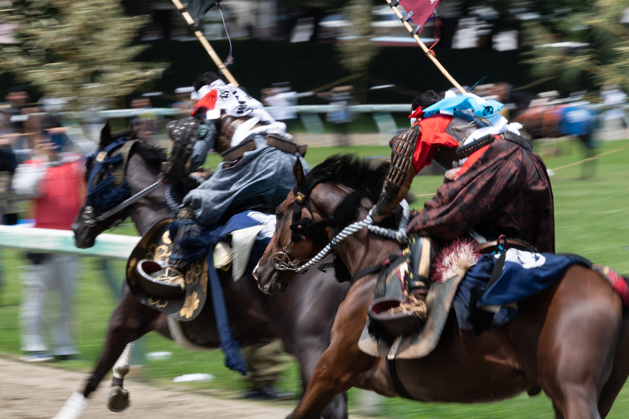 Horse riders in suits of armor charge on horseback during a jousting event, with spectators in the blurred background.