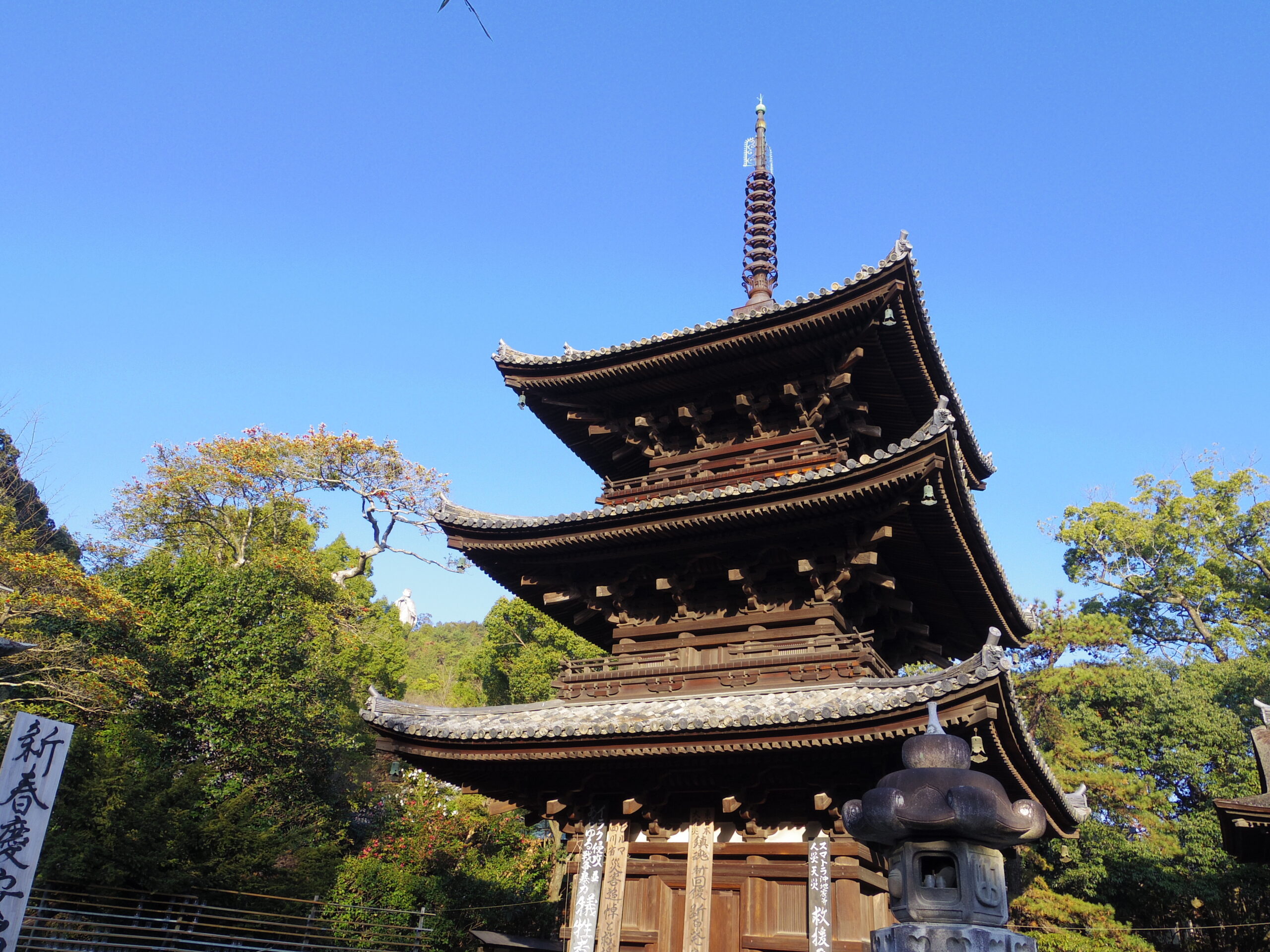 Traditional multi-tier wooden pagoda with curved eaves against a clear blue sky, amid a temple complex and trees in autumn colors.