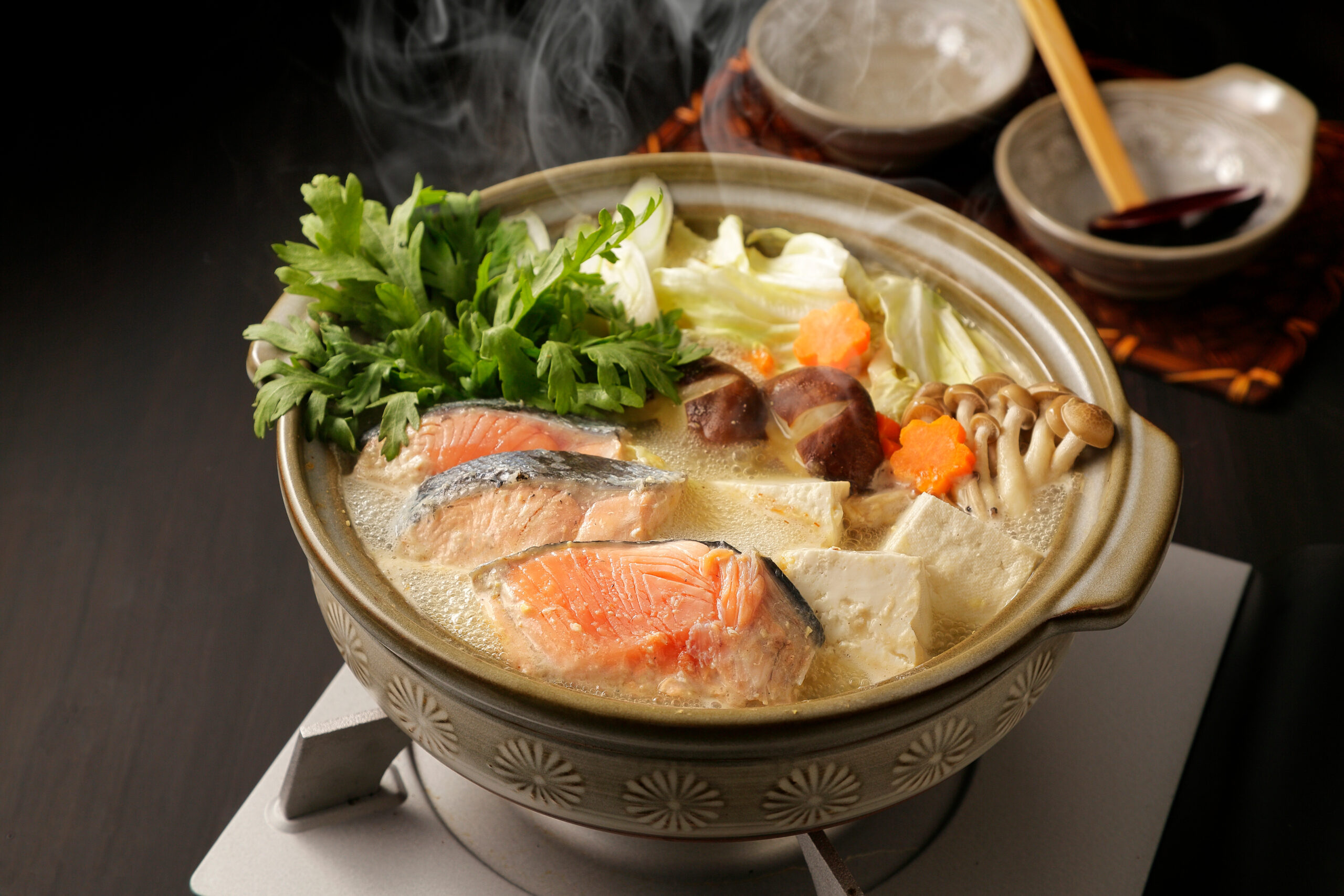 Steaming hot pot filled with salmon slices, tofu, mushrooms, napa cabbage, carrot shapes, and fresh green herbs such as parsley, in a tan ceramic pot.