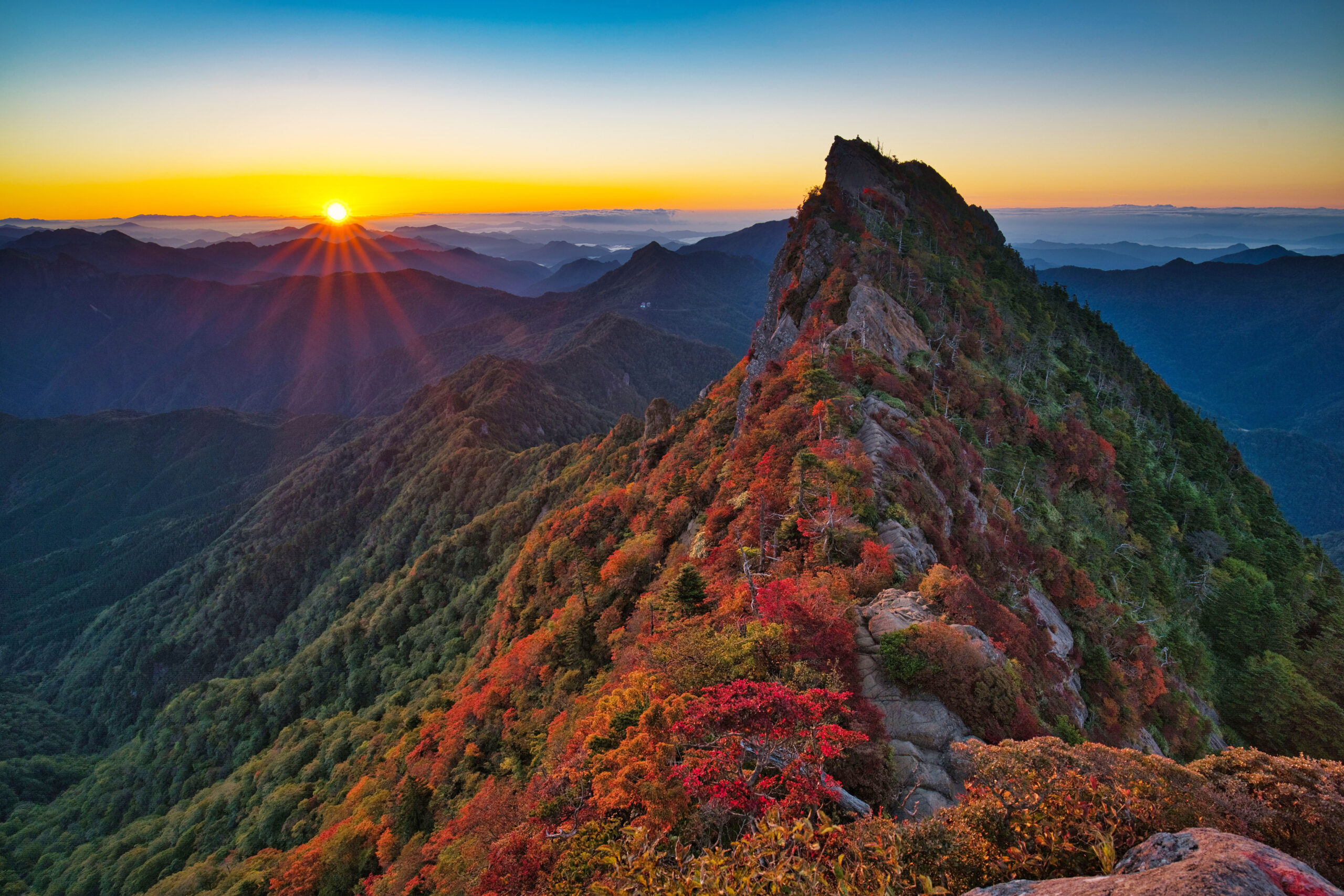 Sunset over a jagged mountain ridge with autumn-colored trees along the slopes at dusk.