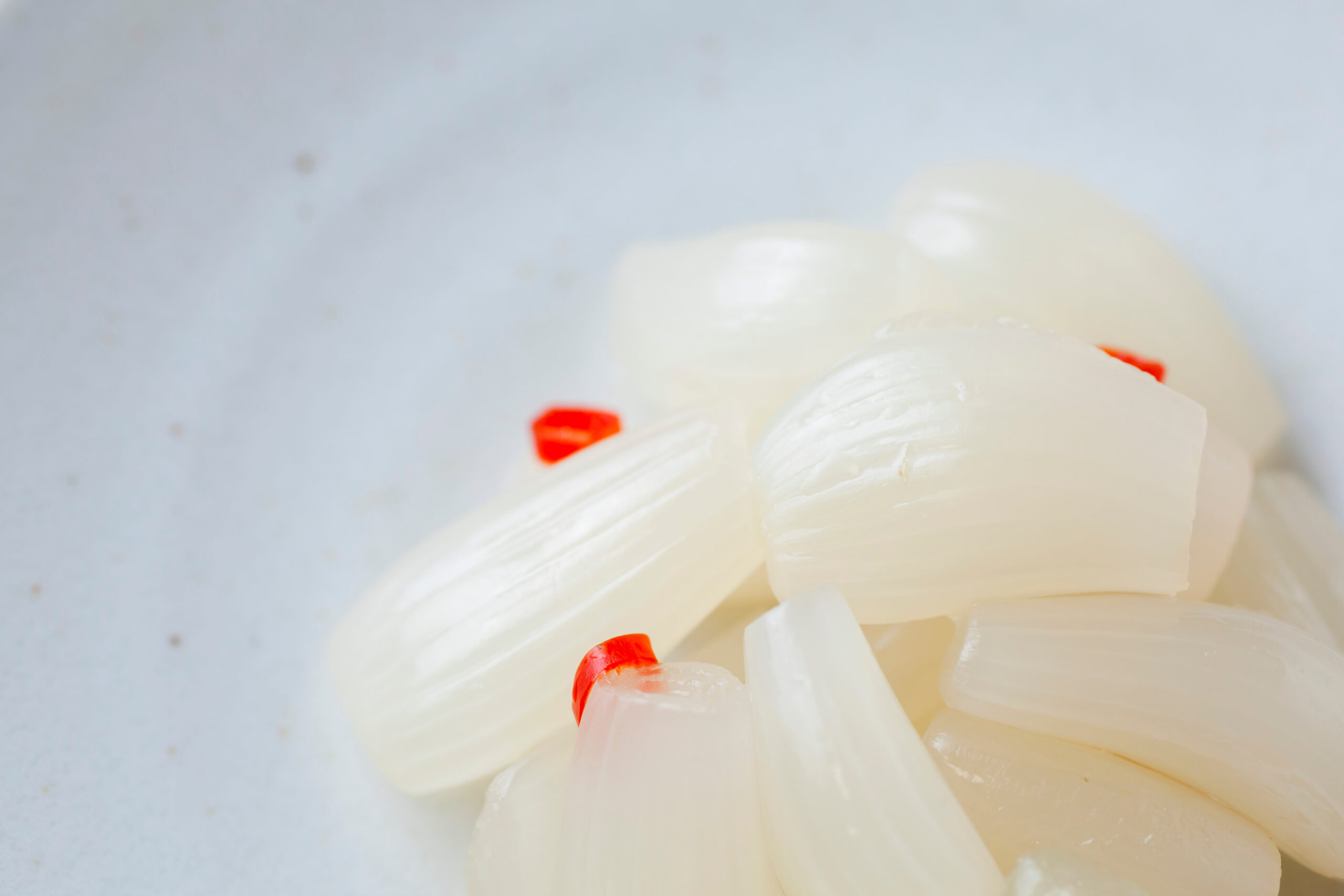 Close-up of peeled white onions with small red pepper pieces on a pale surface.