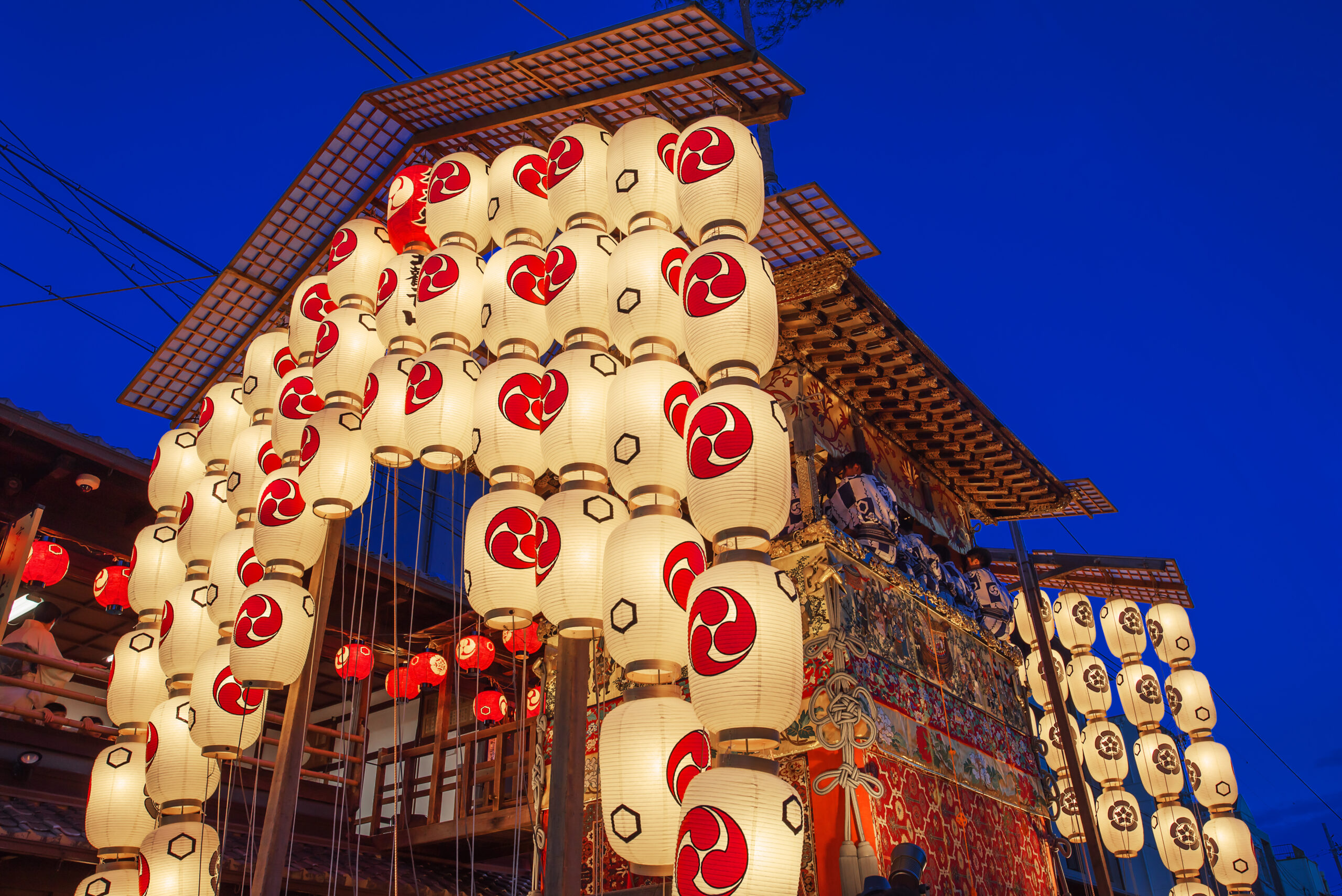 Vertical strings of white lanterns with red symbols, lit for a festival against a deep blue evening sky in a wooden stall setting.