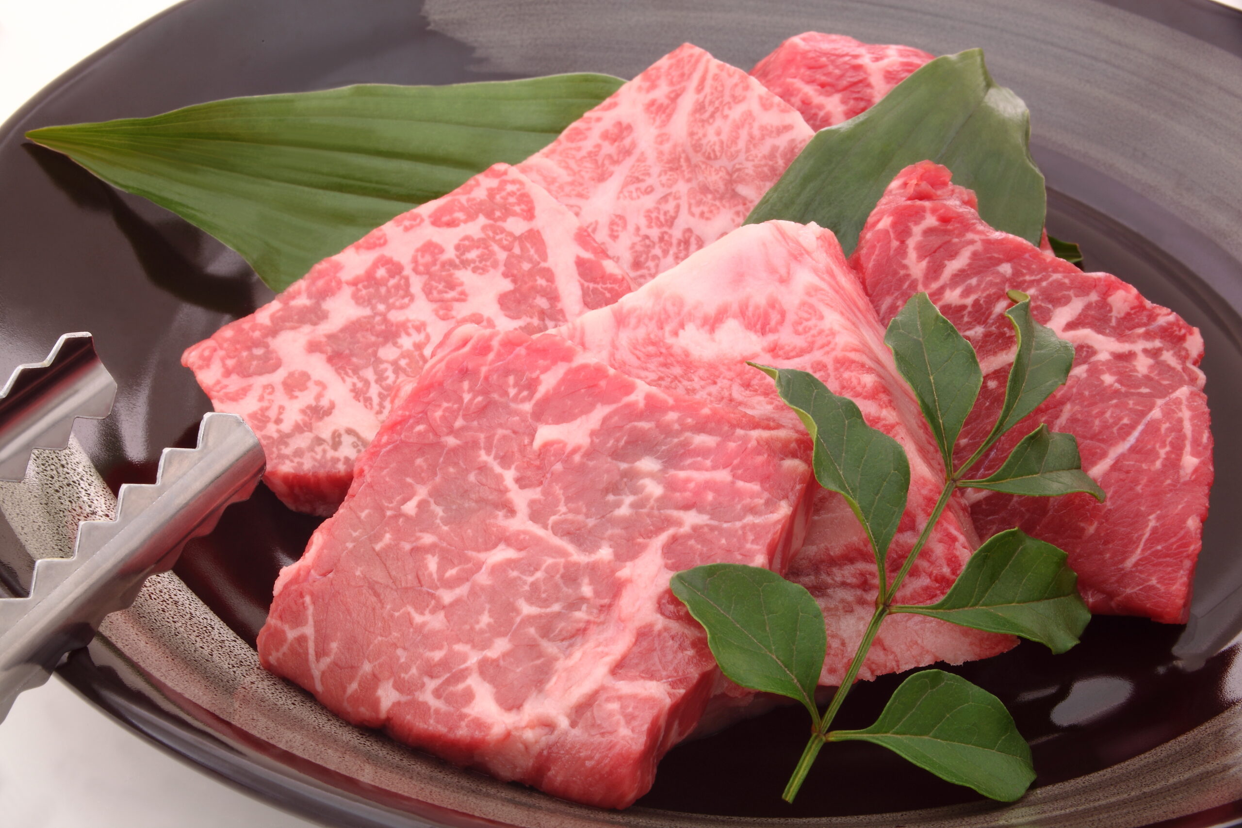 Slices of marbled raw beef with green herbs on a dark plate and metal tongs nearby