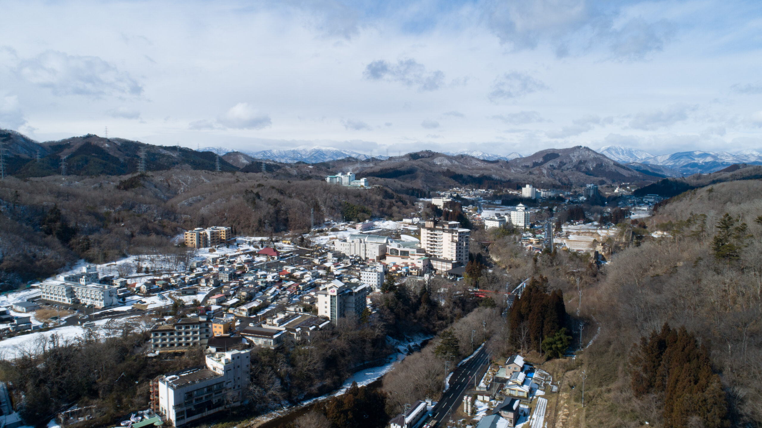A snowy town nestled in a valley with green hills and distant snow-capped mountains under a cloudy blue sky.