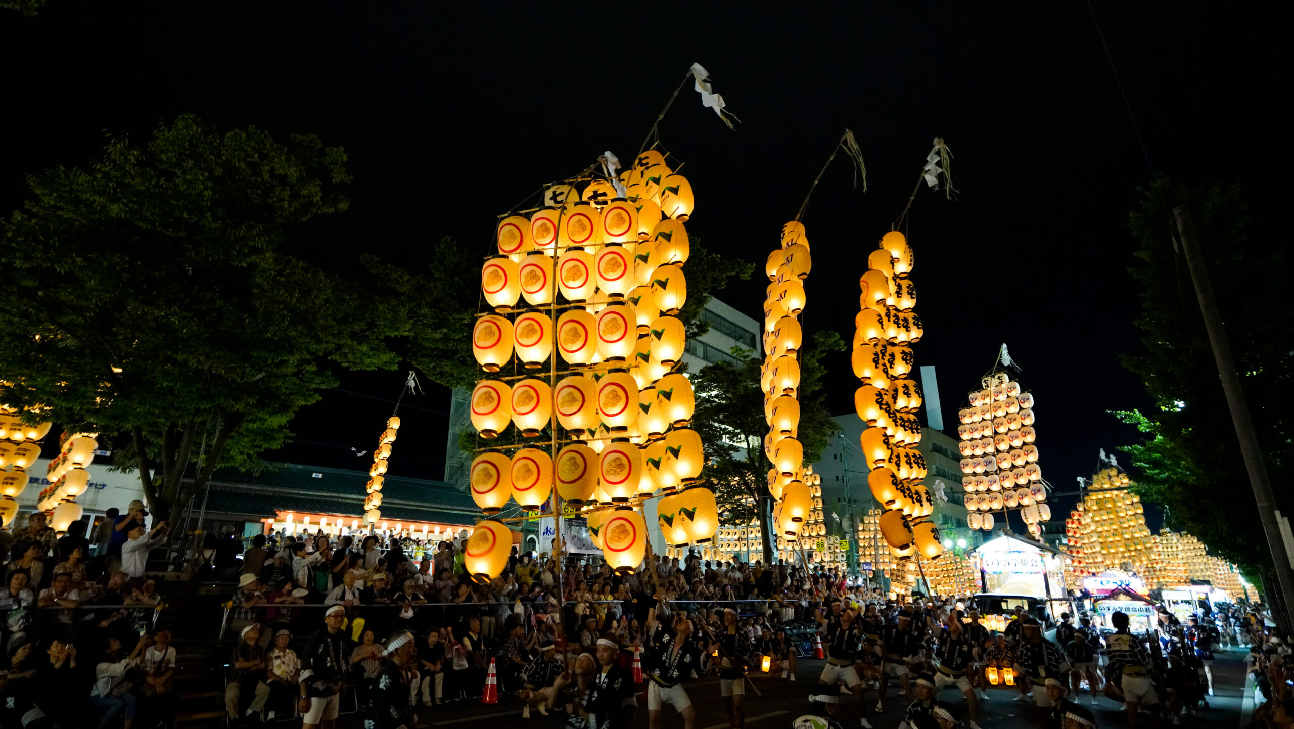 Night festival street scene with tall illuminated yellow lanterns on bamboo frames and a large crowd gathered at night