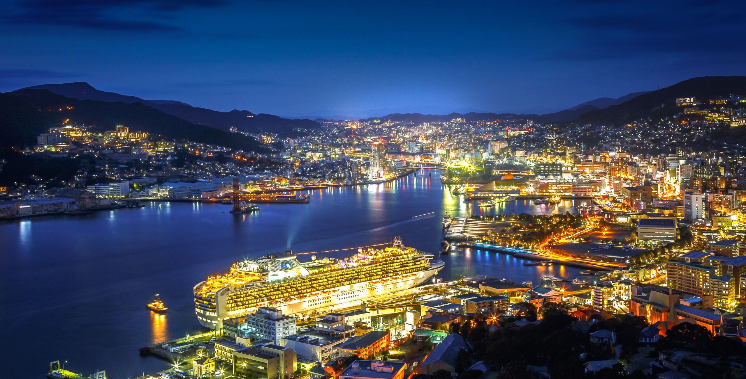 Nighttime cityscape of a harbor with a large illuminated cruise ship docked along the waterfront