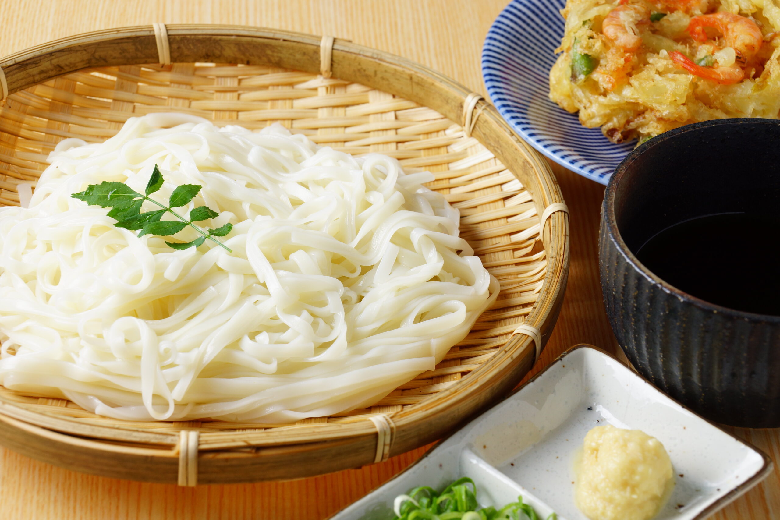 White udon noodles in a woven bamboo basket with dipping sauces and a plate of vegetable tempura nearby.