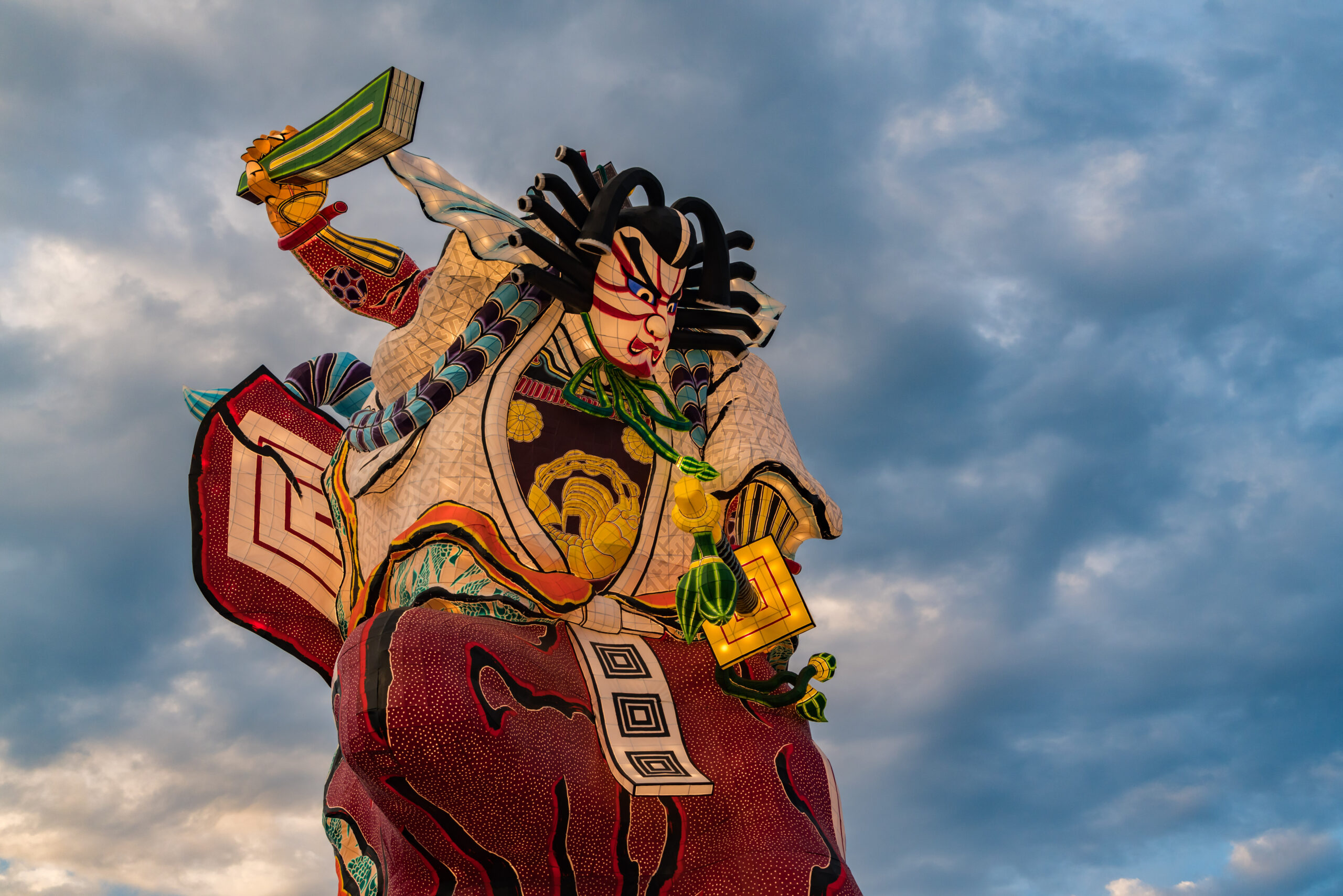 Colorful dragon lantern sculpture rising into a cloudy blue sky, with red, gold, and turquoise patterns and geometric details.