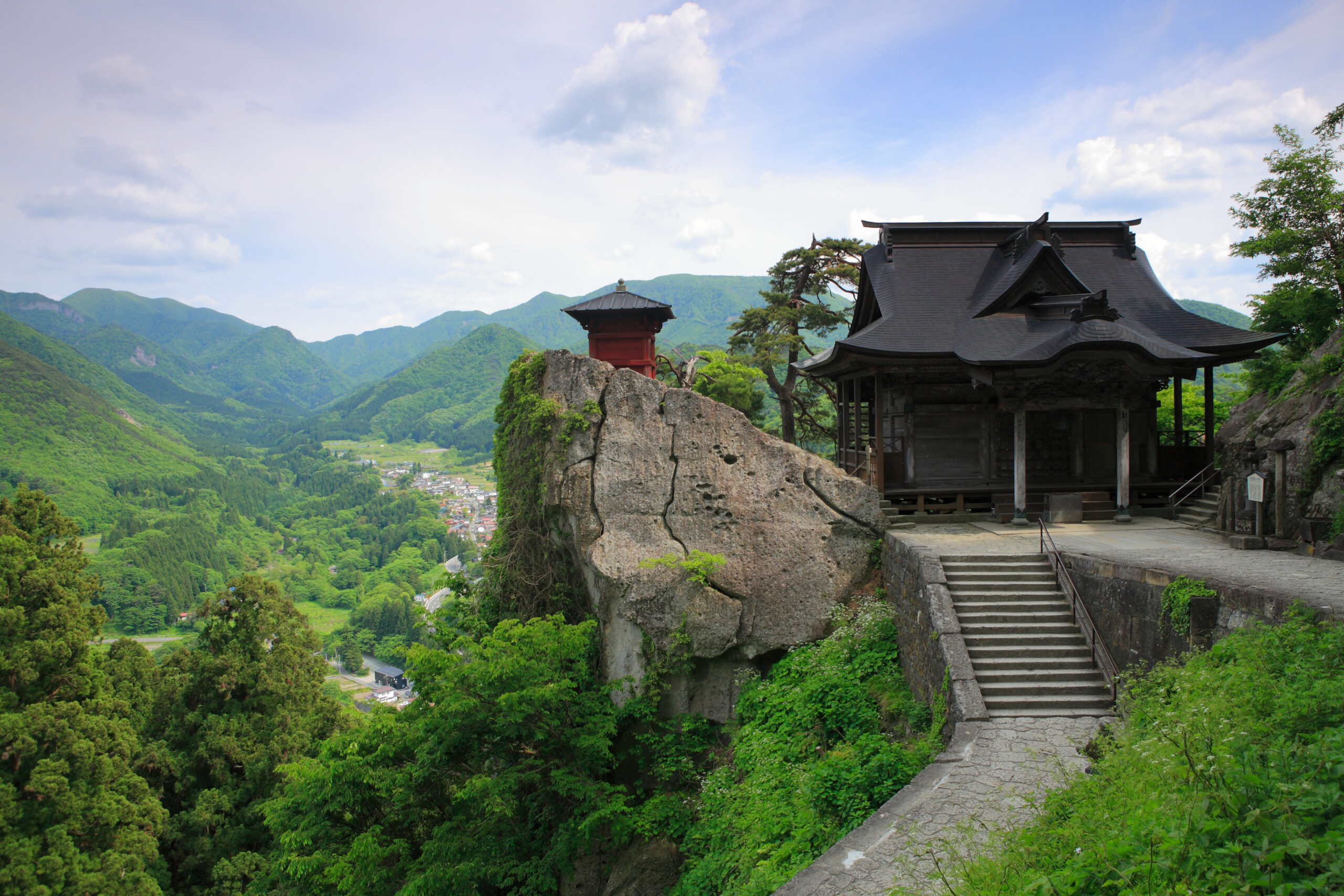 Traditional Japanese temple on a rocky cliff with stairs, overlooking a green valley and distant mountains.