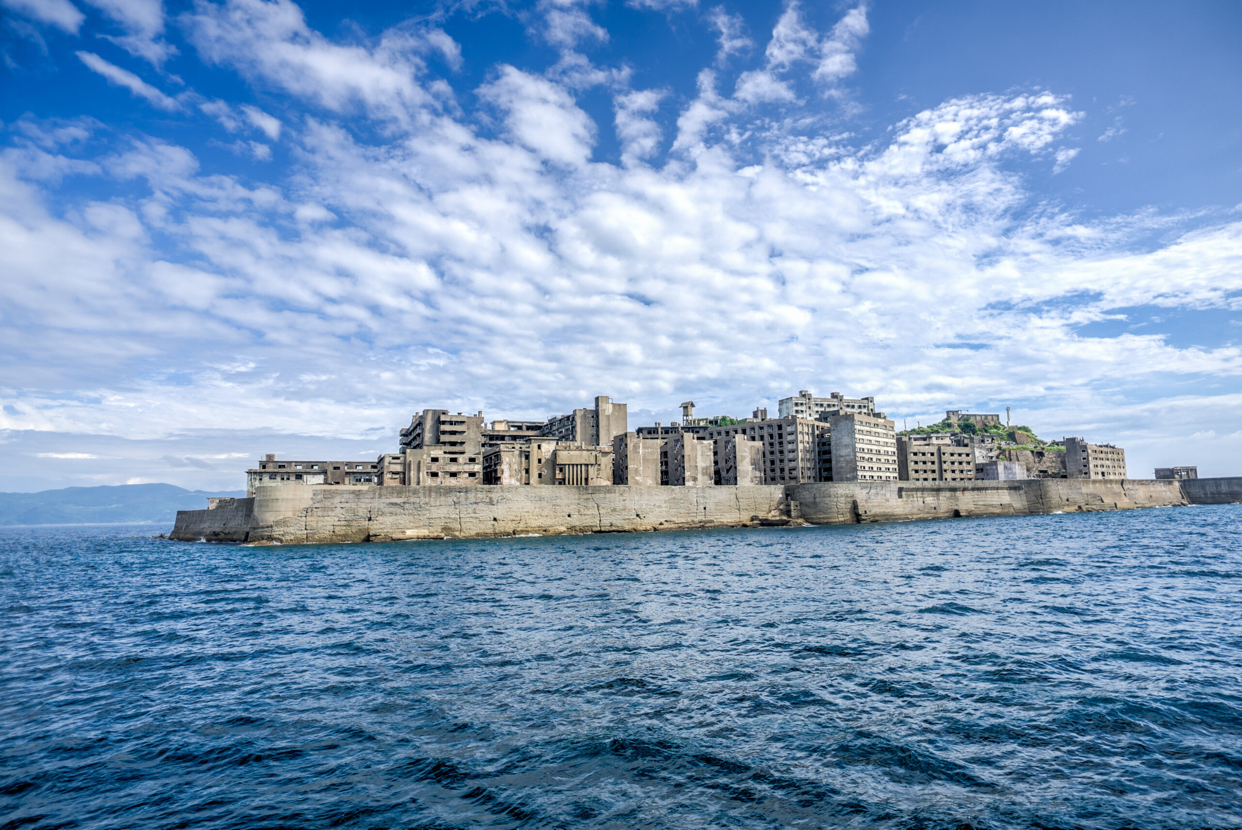 Abandoned concrete waterfront buildings along a seawall under a bright blue sky with scattered clouds.