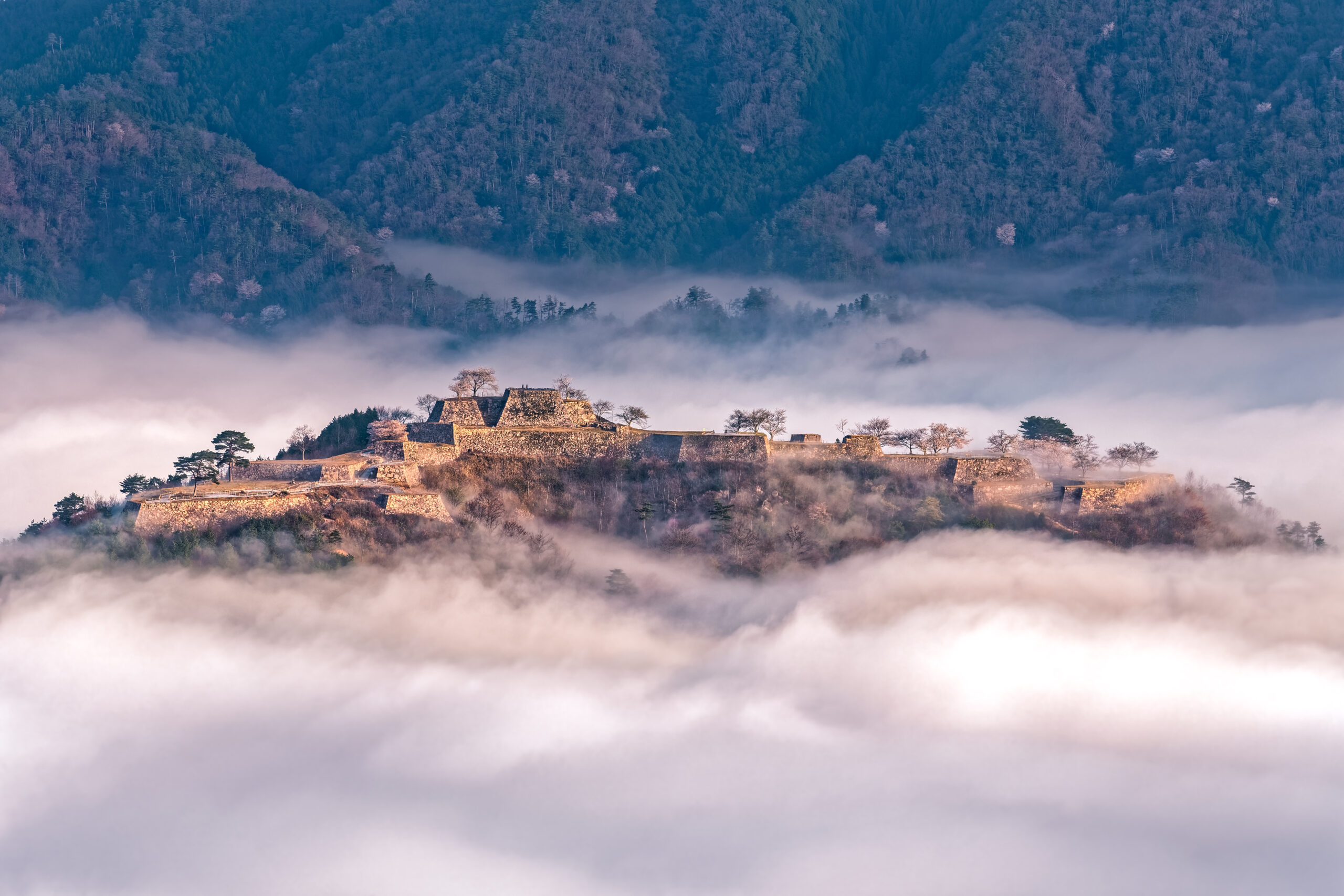 Stone fortress perched on a hill above fog with forested mountains in the background