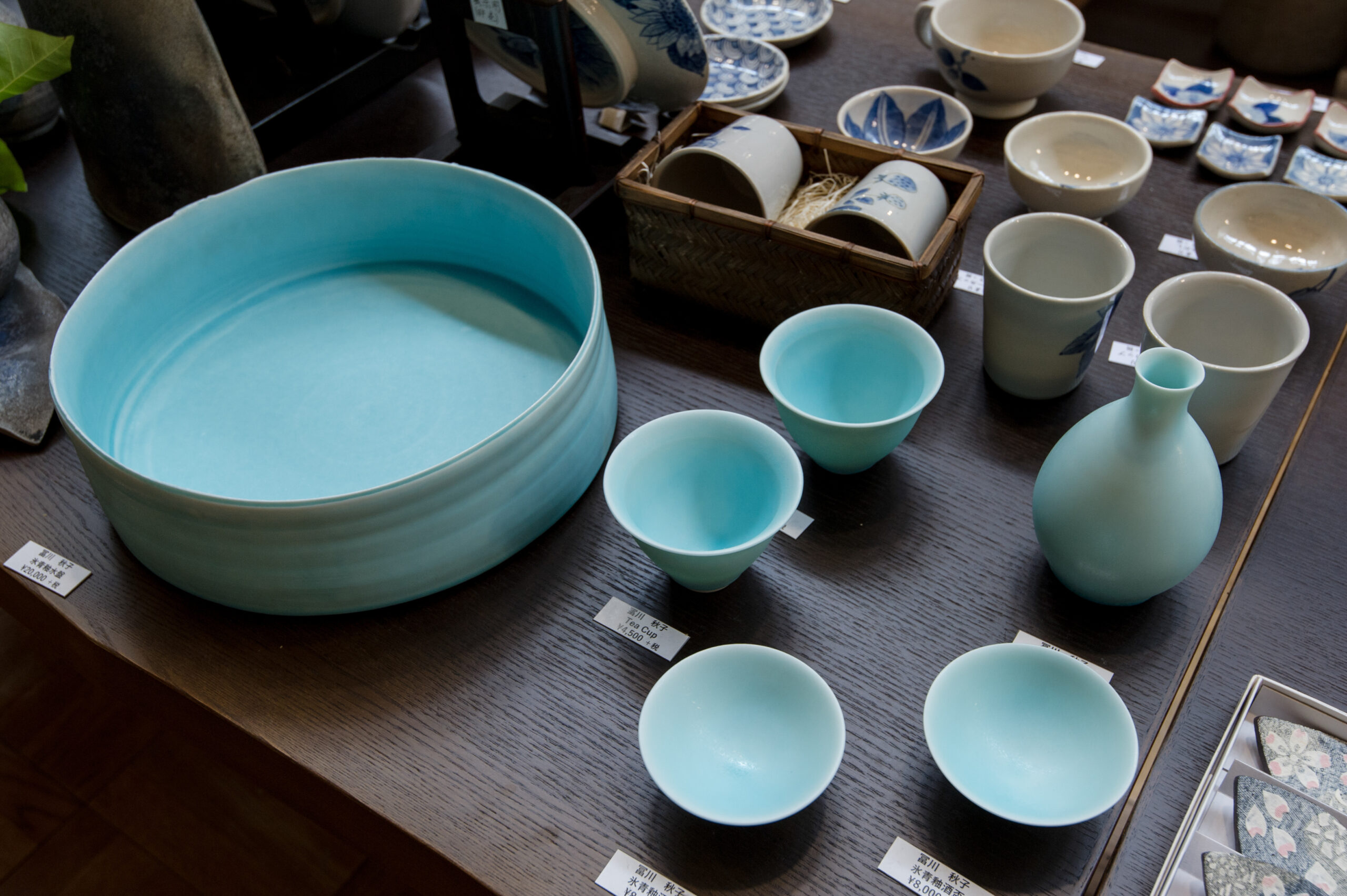 Display of blue-green ceramic bowls and cups on a wooden table, with price tags and a woven basket of pottery pieces in the background