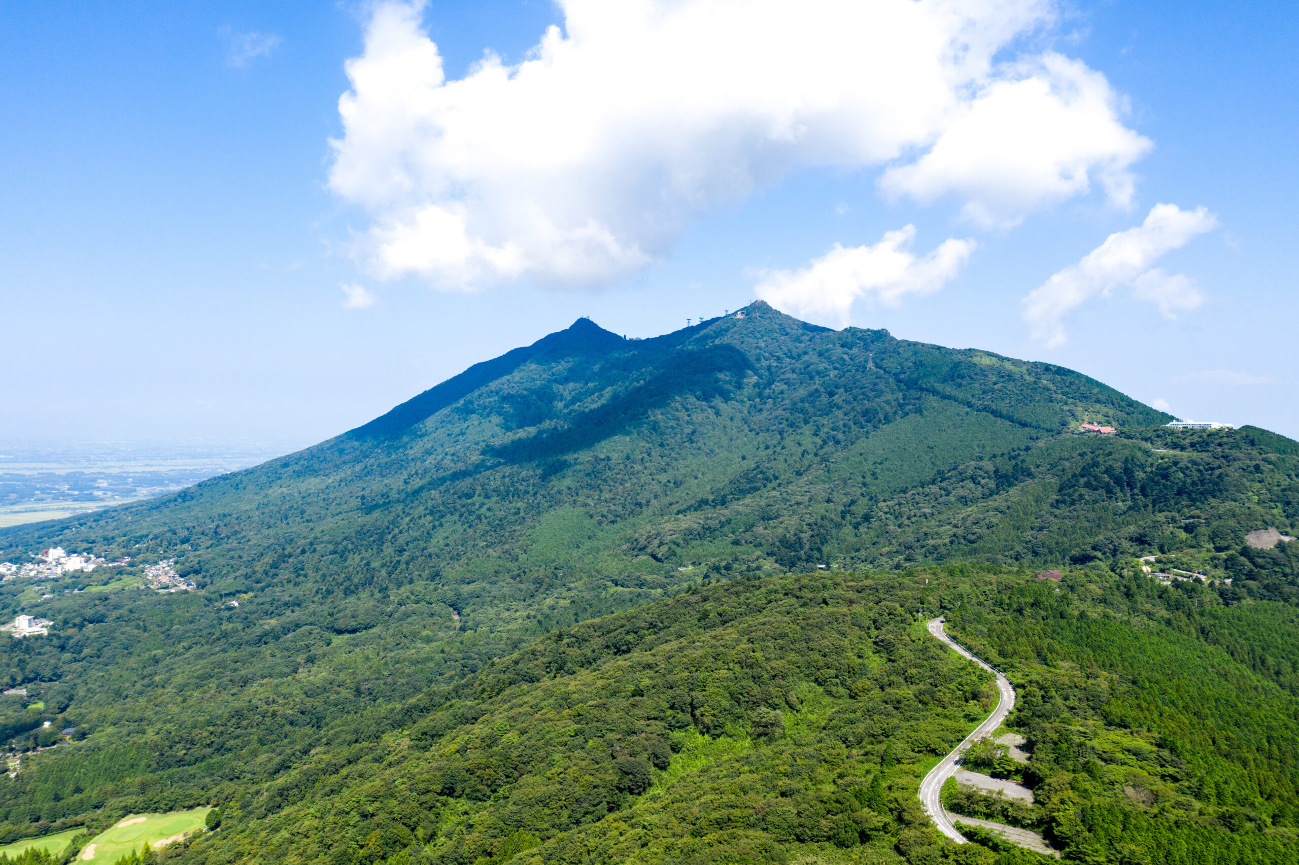 Winding road curves along a forested hillside under a bright blue sky with white clouds, seen from above.