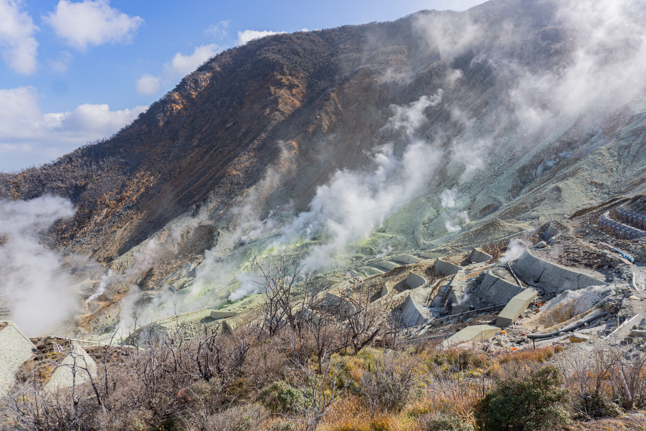 Steep rocky mountainside with rising steam and clouds near a volcanic crater, with concrete blocks and pipes visible on the right.