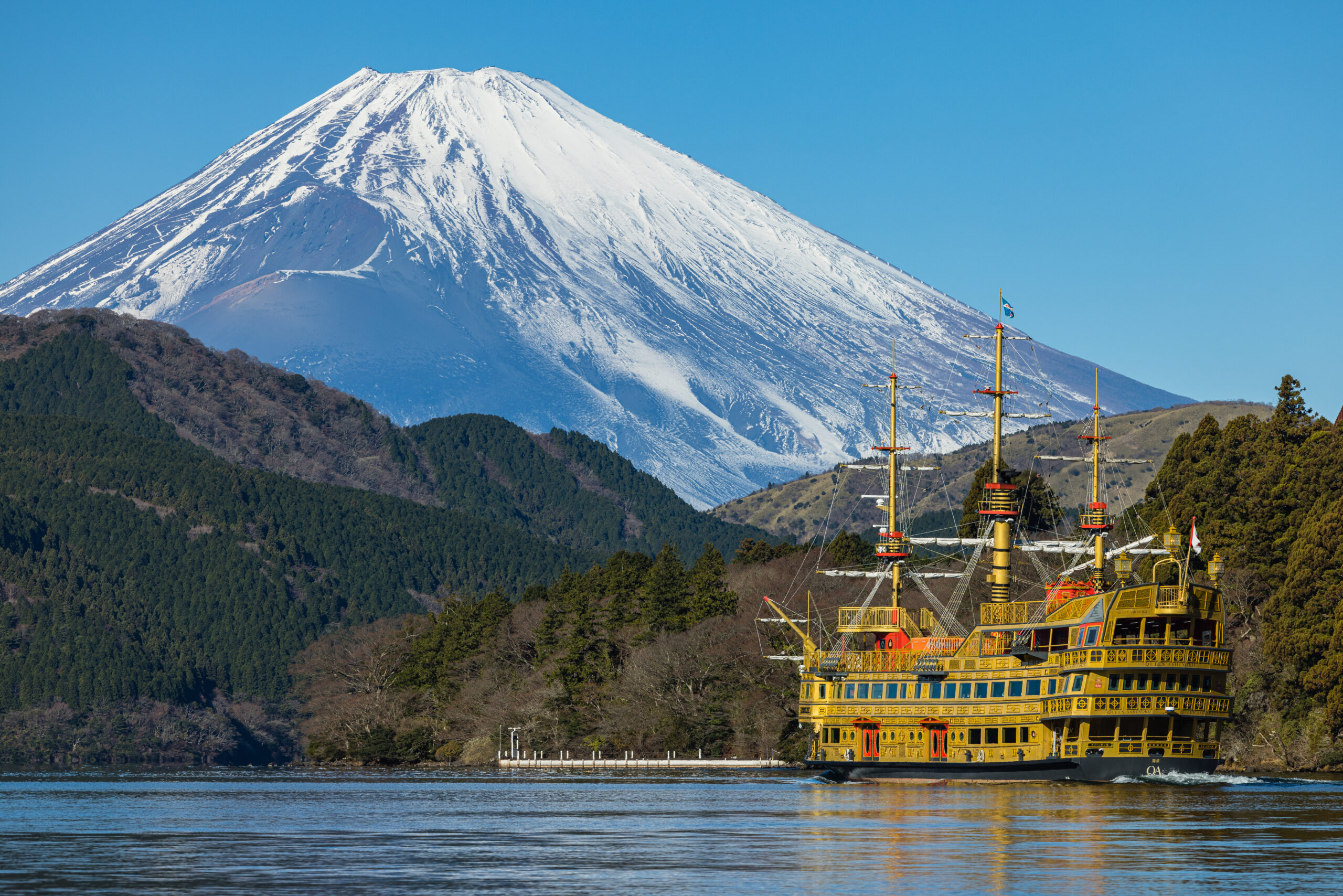Bright yellow, multi-deck ship with tall masts near a forested coastline and a snow-covered mountain in the distance.