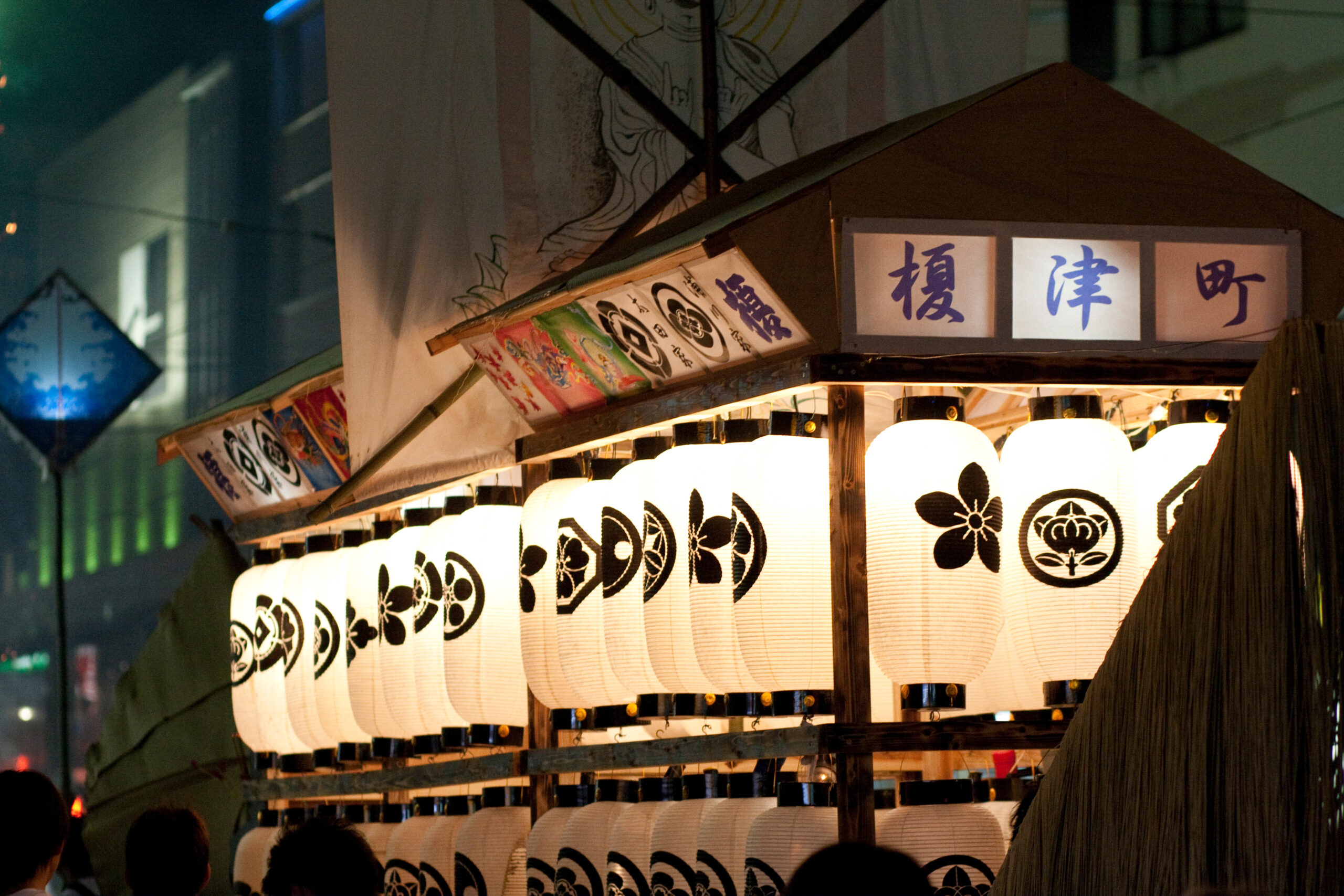 Nighttime festival stall with rows of glowing white paper lanterns hanging on a wooden frame, blue kanji panels on the roof, and colorful banners along the side