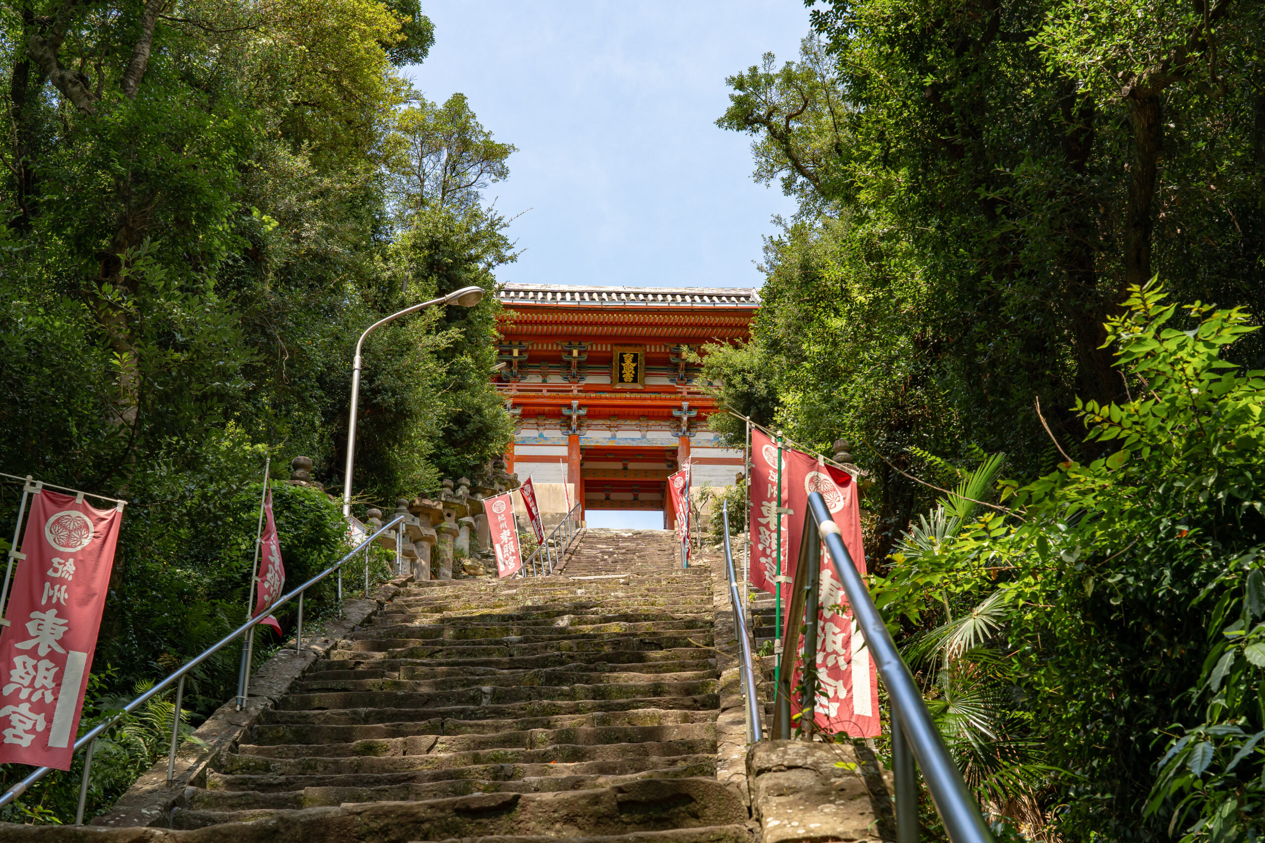 Stone stairs lined with red banners lead up to a traditional temple gate framed by trees and blue sky beneath.