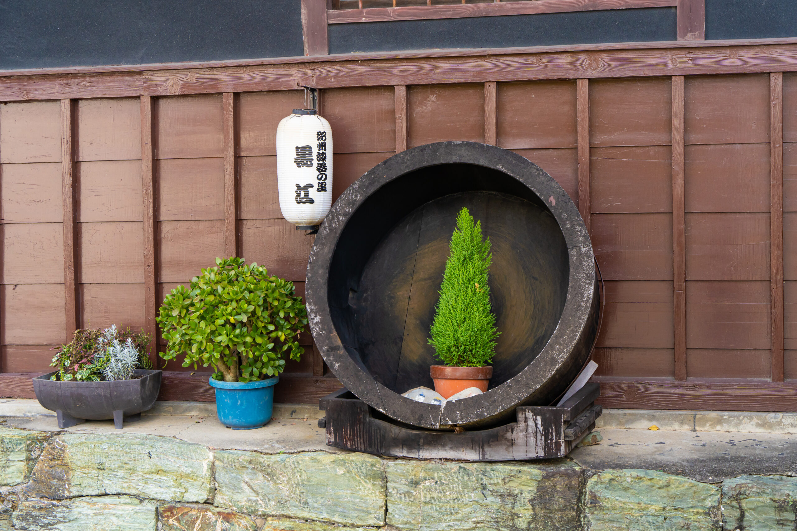 White Japanese lantern hanging on a brown wooden wall above a circular dark tunnel or pipe, with various potted plants in front.