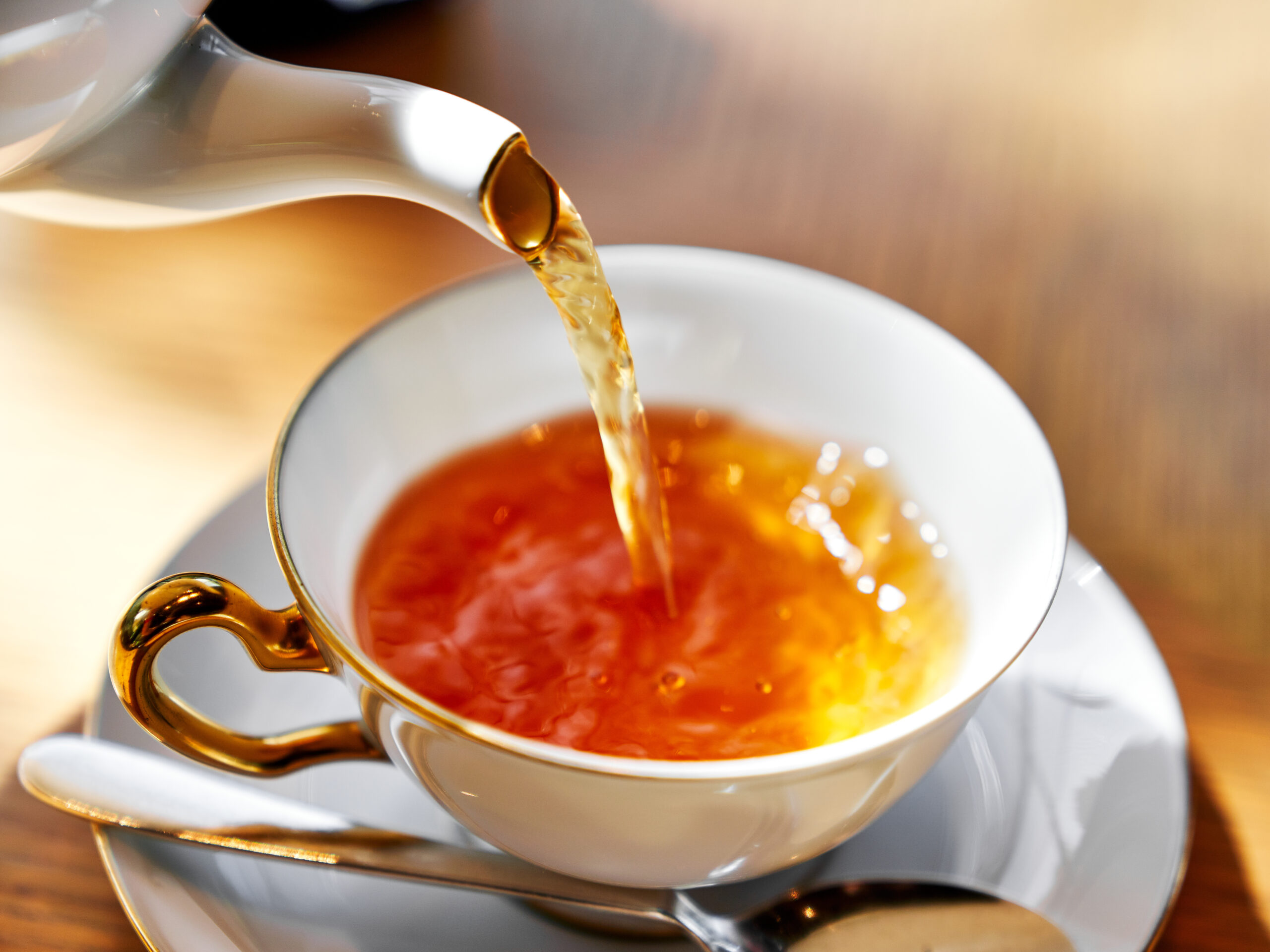 Golden-rimmed teacup and saucer as amber tea is poured from a teapot into the cup, close-up view with warm lighting.