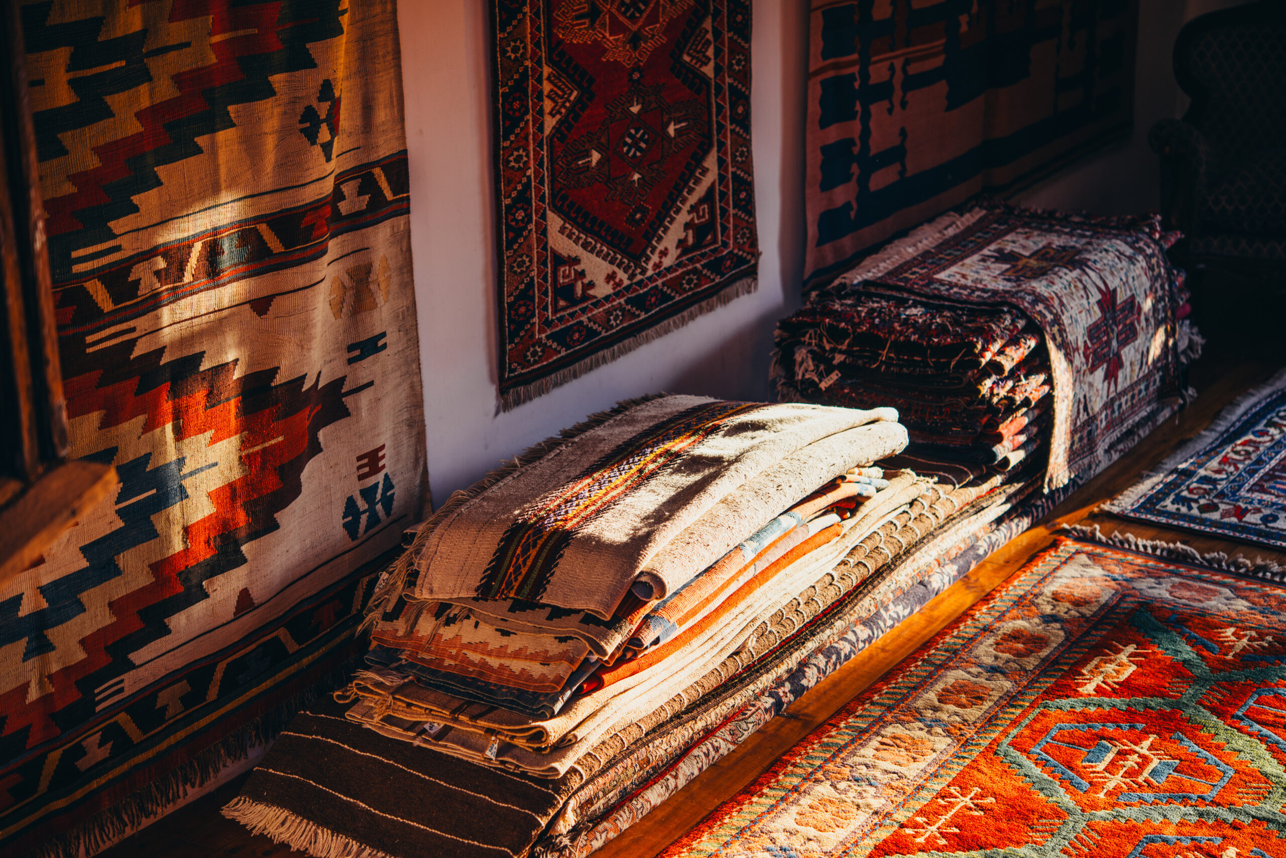 A display of colorful woven rugs and carpets stacked along a wall and on the floor, showing intricate geometric patterns and fringed edges.