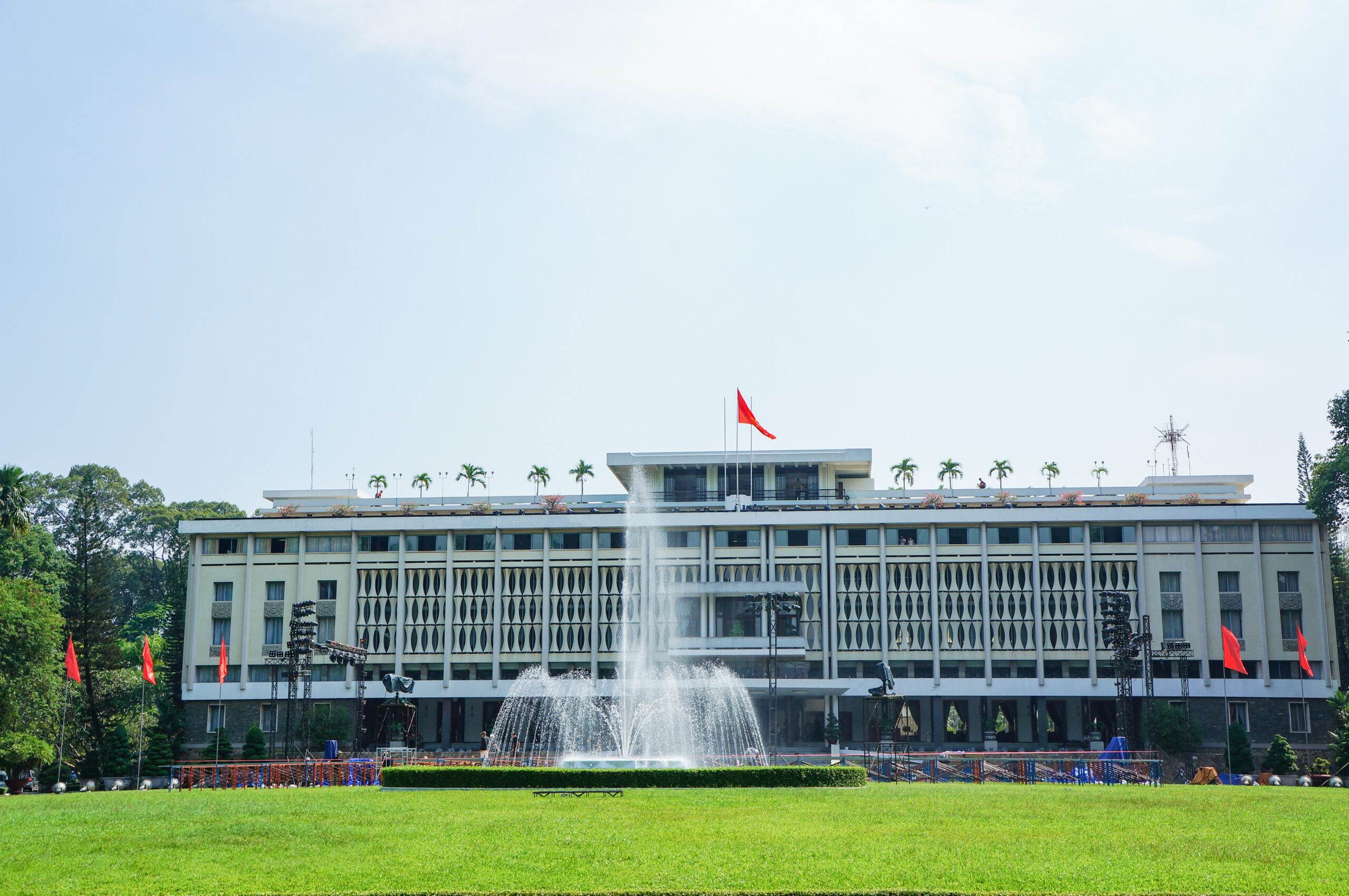 Modern multi-story building with a large fountain in front, red flags along the plaza, and a green lawn beside it.