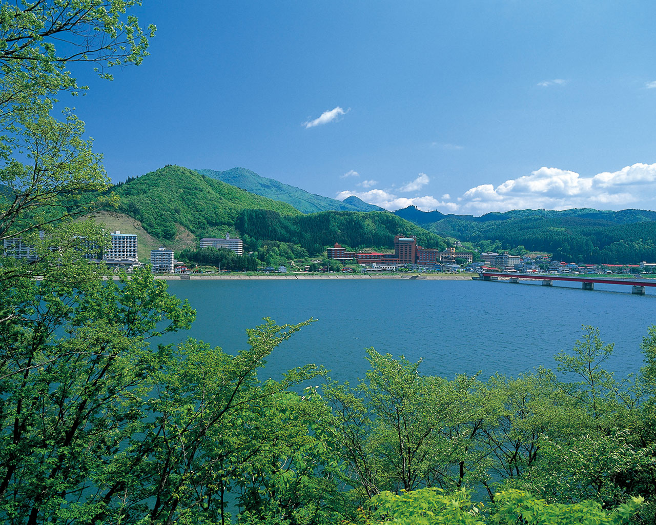A lake with a town along the shore, green hills in the distance and a bridge spanning the water, framed by leafy foreground trees.