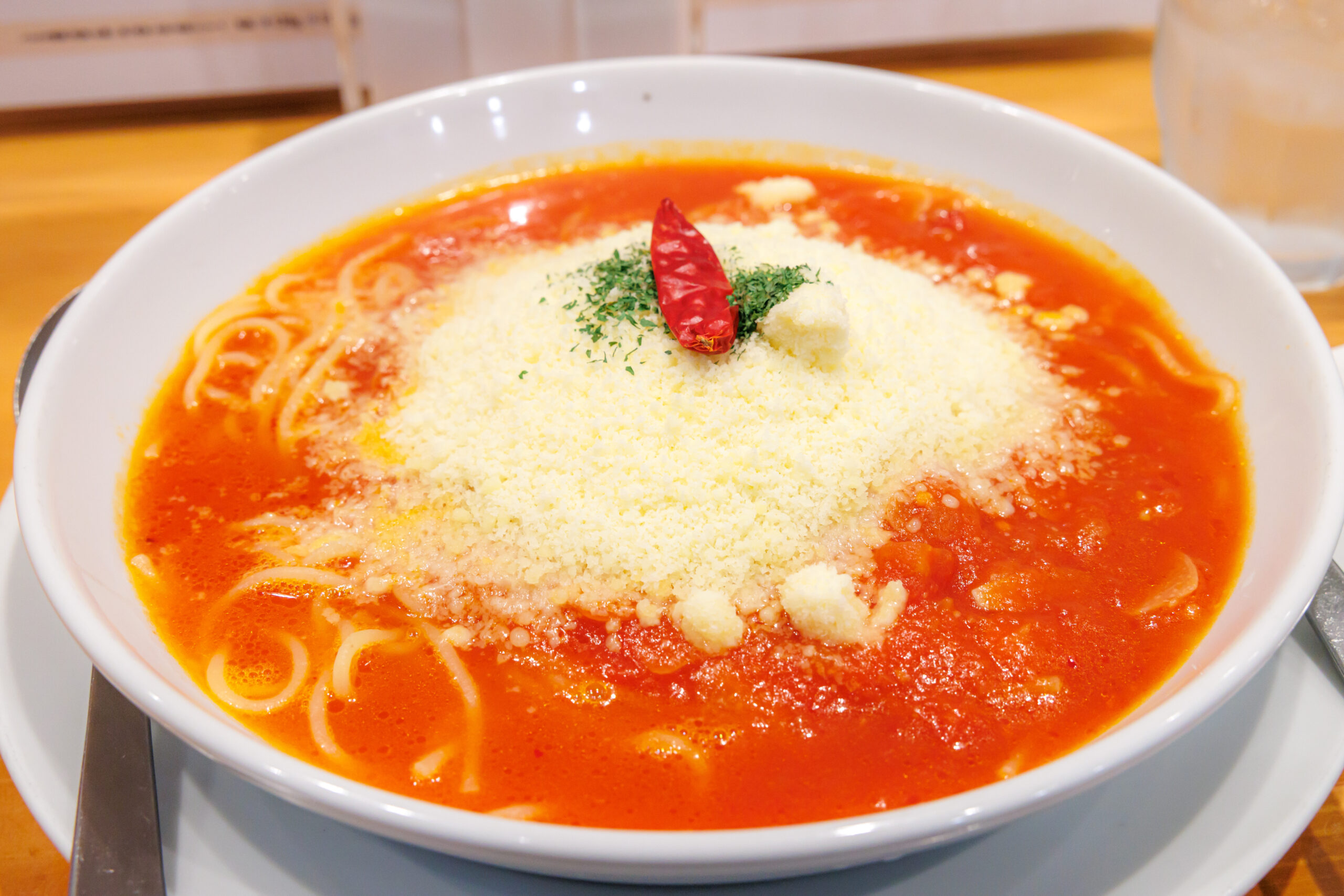 Bowl of tomato-based soup with grated cheese and a single red chili pepper on top, pasta strands visible in the soup.