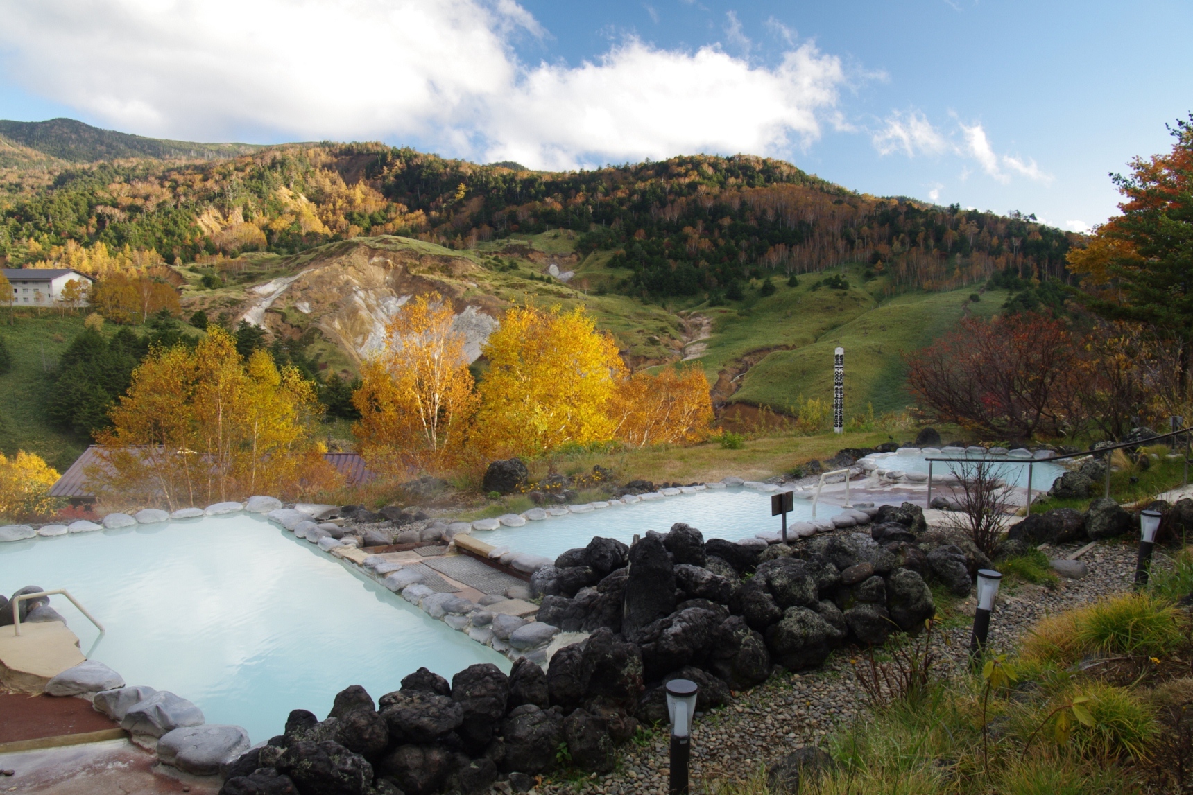 Mountain landscape with autumnal trees surrounding a pale blue hot spring pool bordered by rocks and a gravel path in the foreground, under a bright blue sky with white clouds.