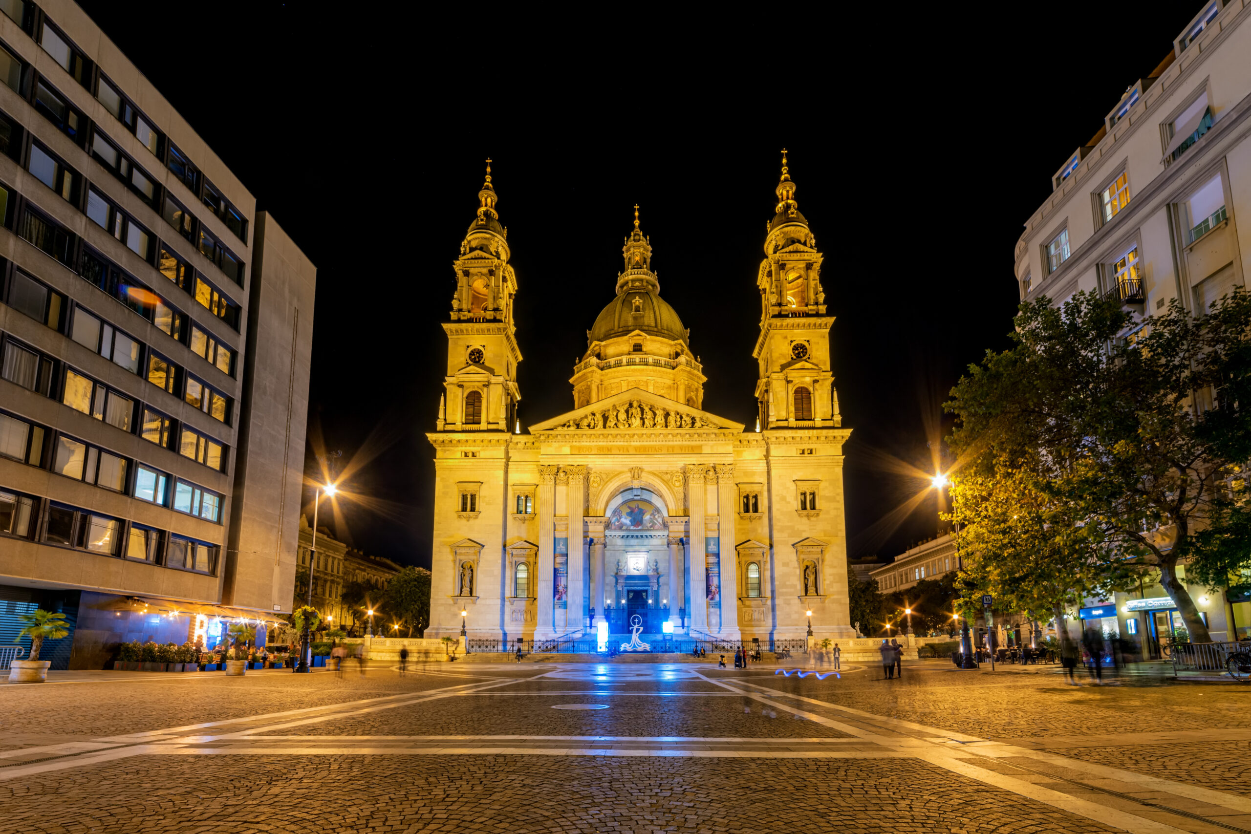 Night view of a grand, illuminated cathedral with a central dome and two towers, set in a city square at night, framed by modern buildings on both sides.