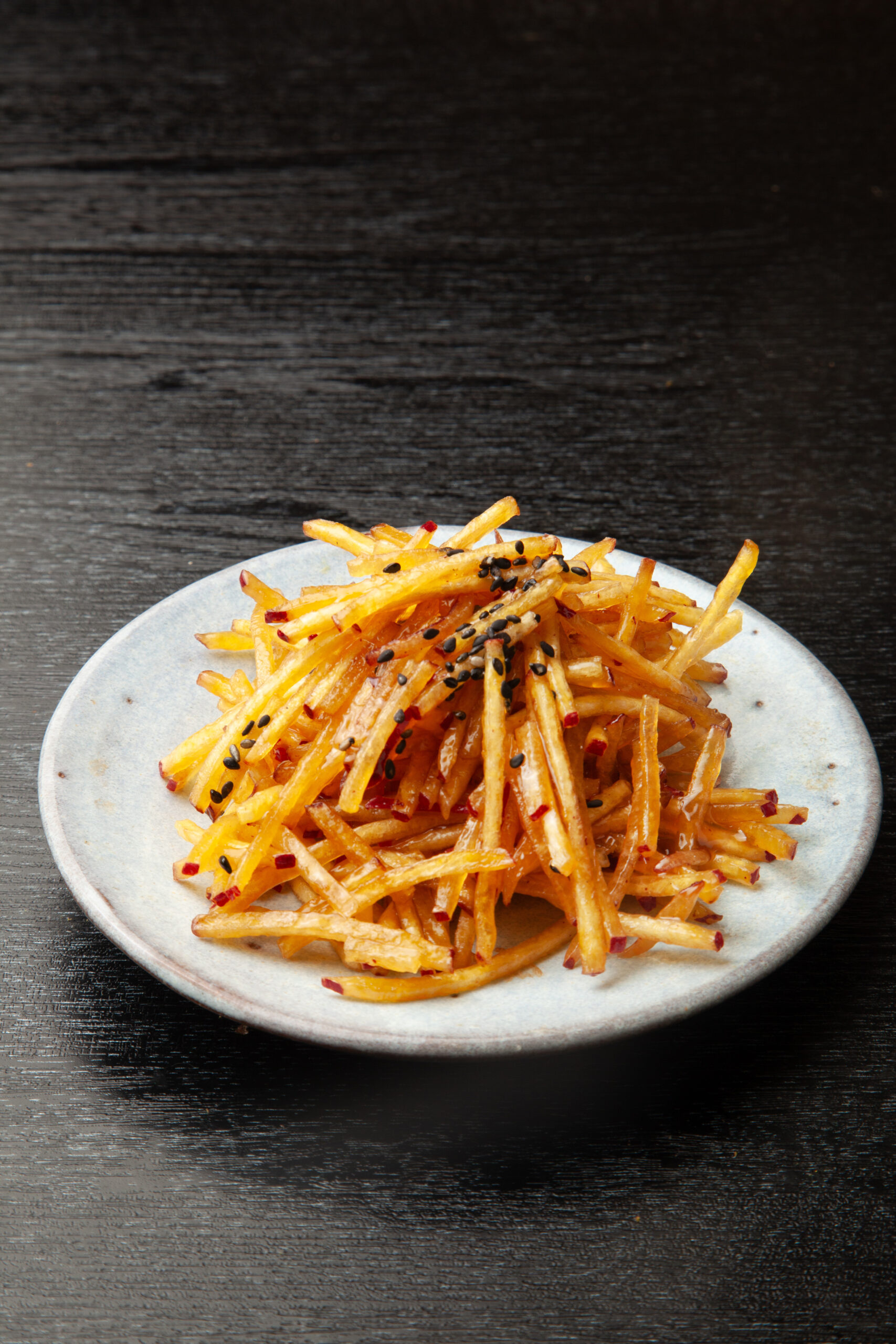 Plate of crispy French fries sprinkled with red pepper flakes and black sesame on a dark wooden table surface.