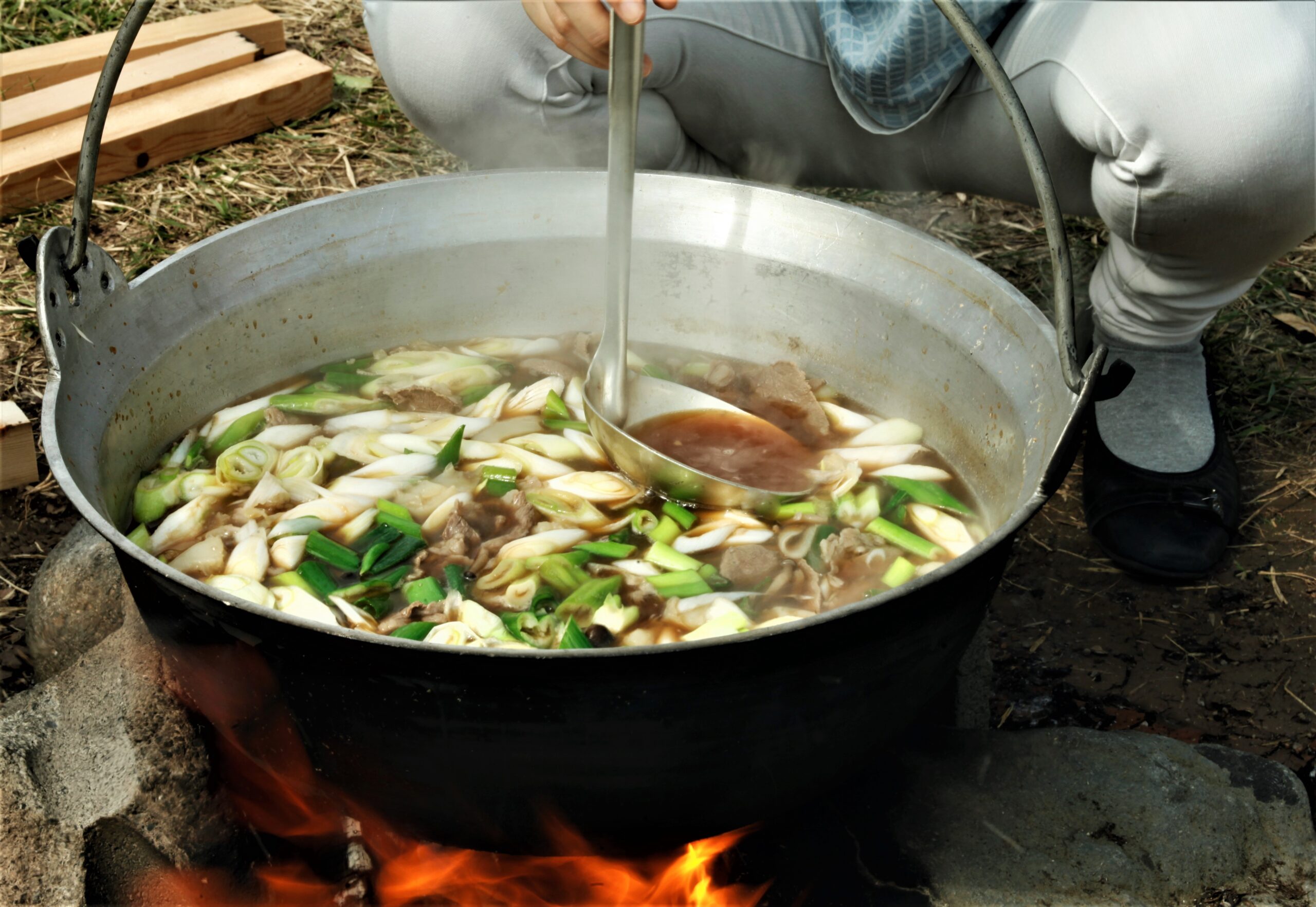 A person stirs a large pot of soup outdoors over an open flame, with vegetables visible in the broth.