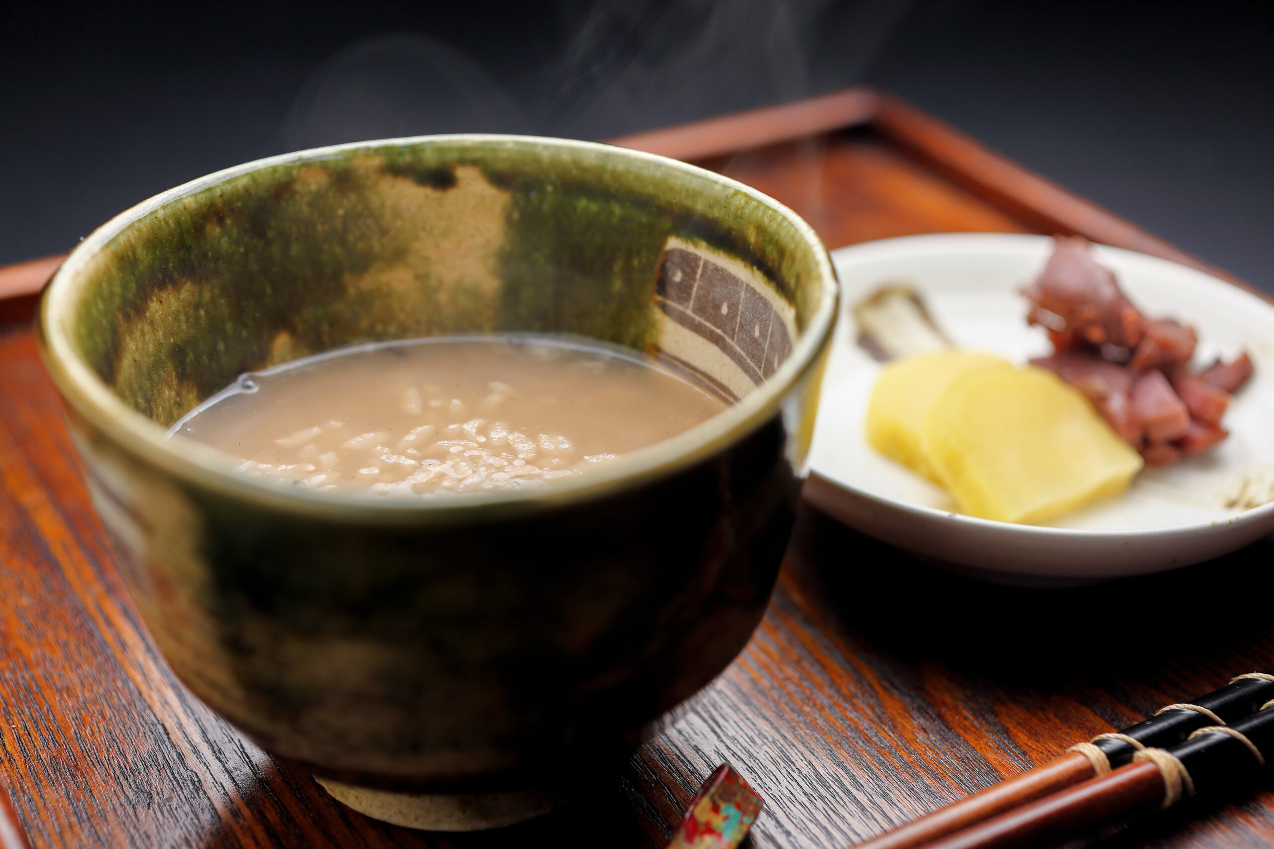 Creamy rice porridge in a green ceramic bowl with steam, on a wooden table, beside chopsticks and a plate of lemon and meat pieces.