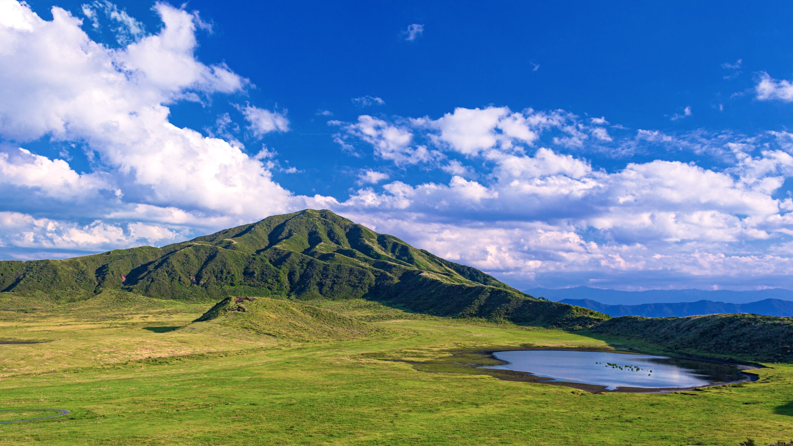 Green rolling hills under a bright blue sky with scattered white clouds and a small pond in the foreground.